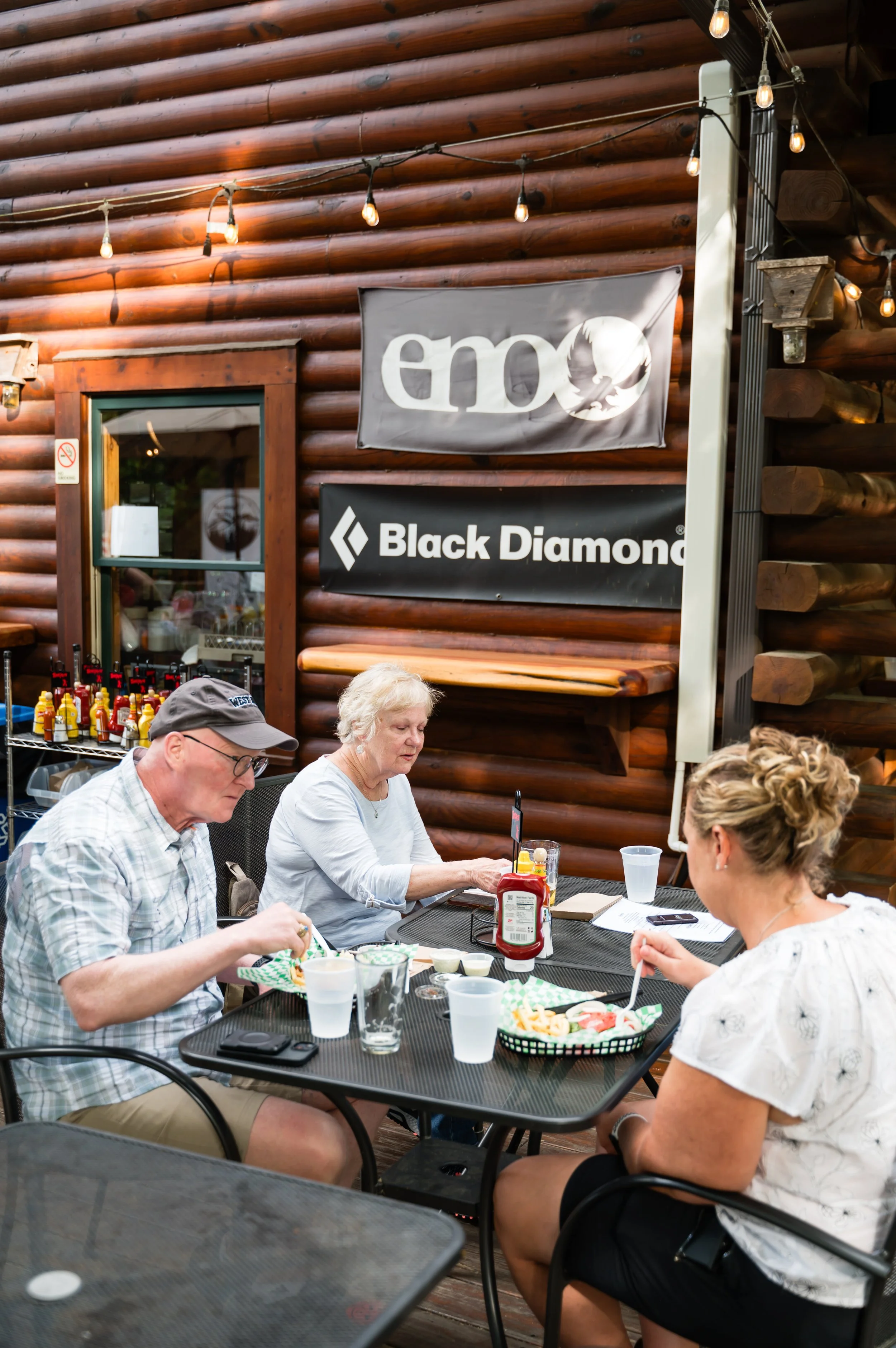 A group of five people sitting at a table outside of a log cabin-style building, eating food and drinking drinks. The building has a wooden log exterior, and there are string lights hanging above them. The table has condiments and a basket of fries.