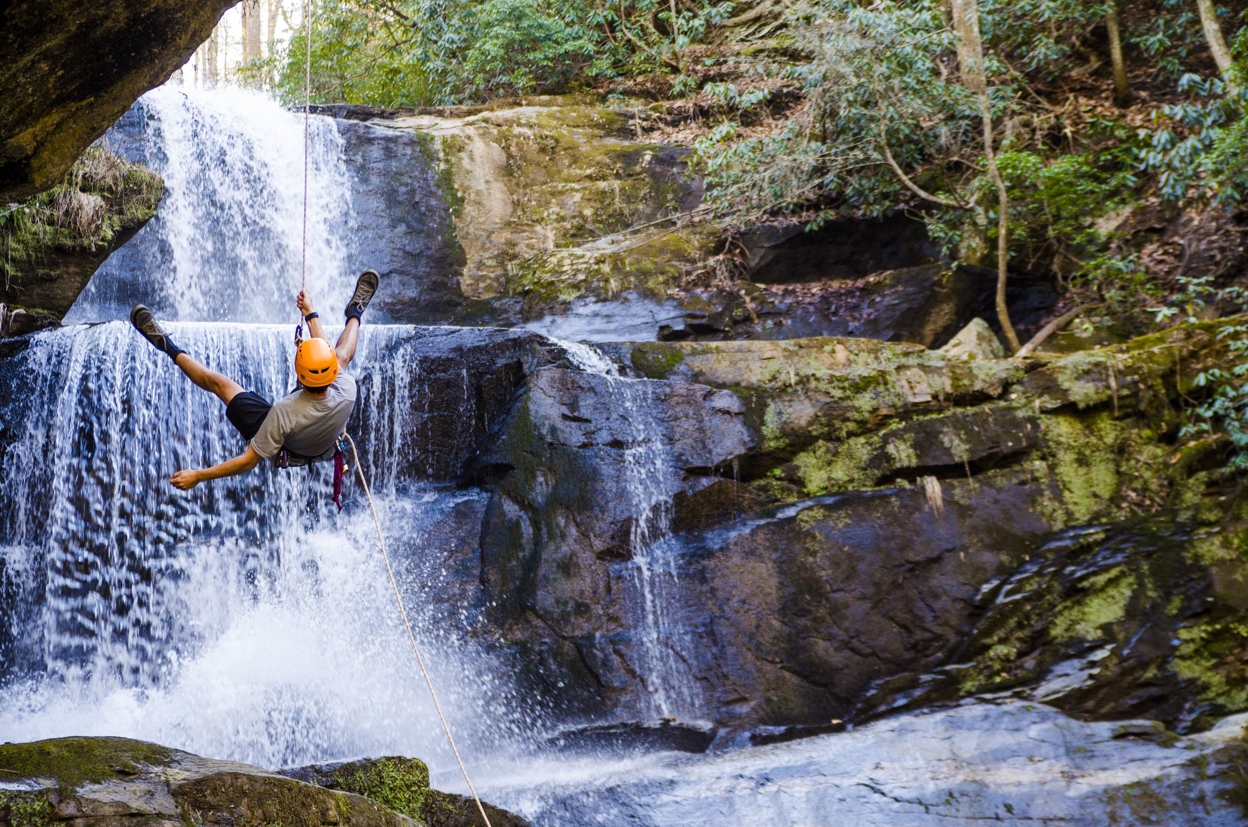A person in an orange helmet and tan shirt rock climbing on a waterfall in a forest.