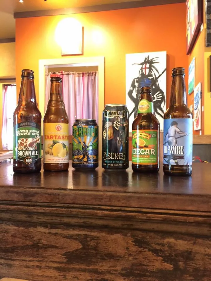 Six bottles and cans of beer and soda arranged on a wooden surface in front of an orange wall. The labels on the bottles include Brown Ale, Lemon Ginger, a plant-themed soda, Black Label Beer, and an ale with a woman on the label.