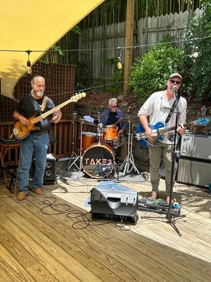 Three musicians performing outdoors on a wooden deck, with a man singing and playing guitar, a man on drums, and a man playing bass guitar, surrounded by greenery and string lights.