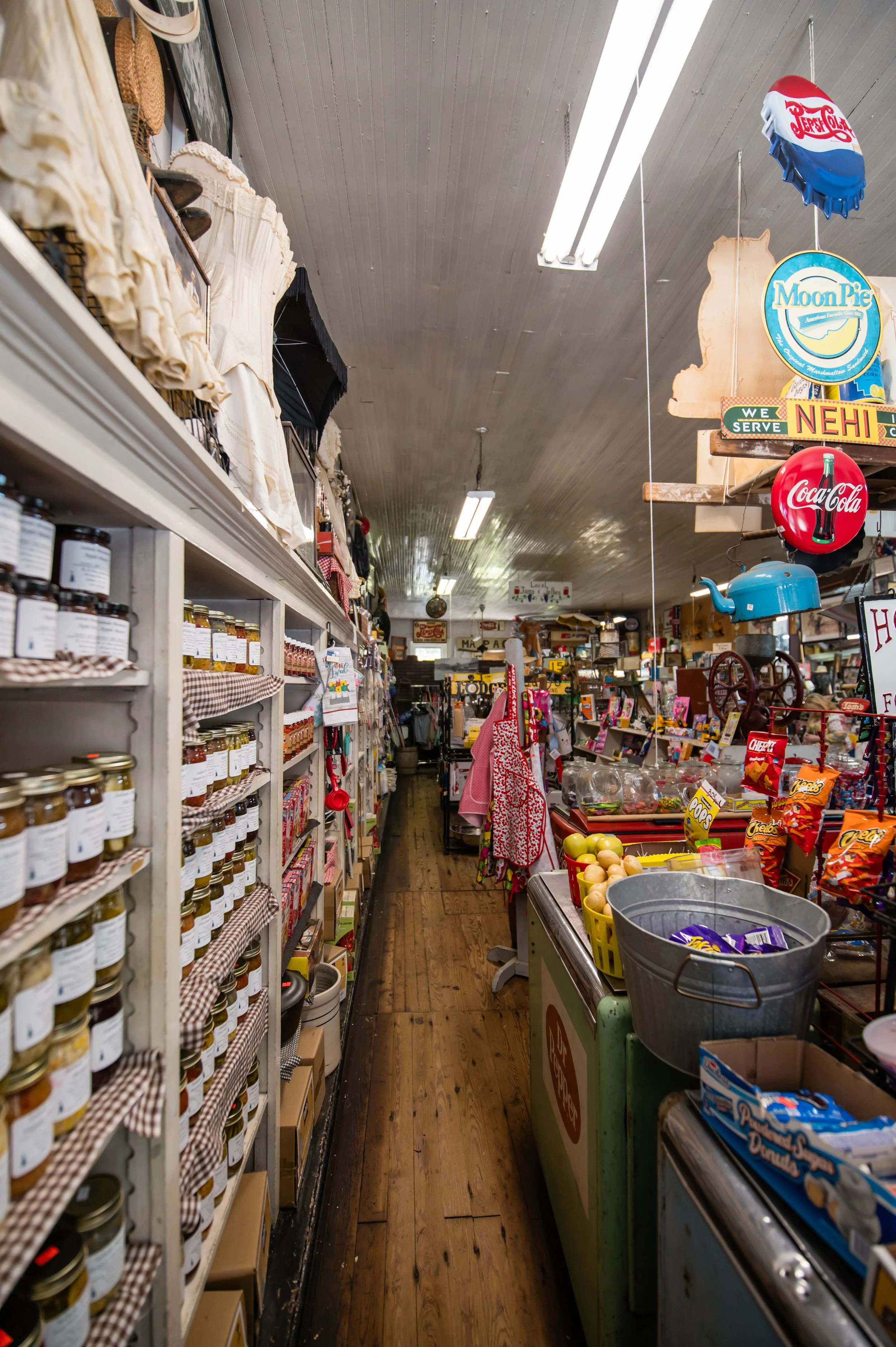 View of an aisle inside a grocery or general store, with shelves on the left stocked with jars and bottles, and various products on the right including a cart with produce, signs, and decorative items hanging from the ceiling.