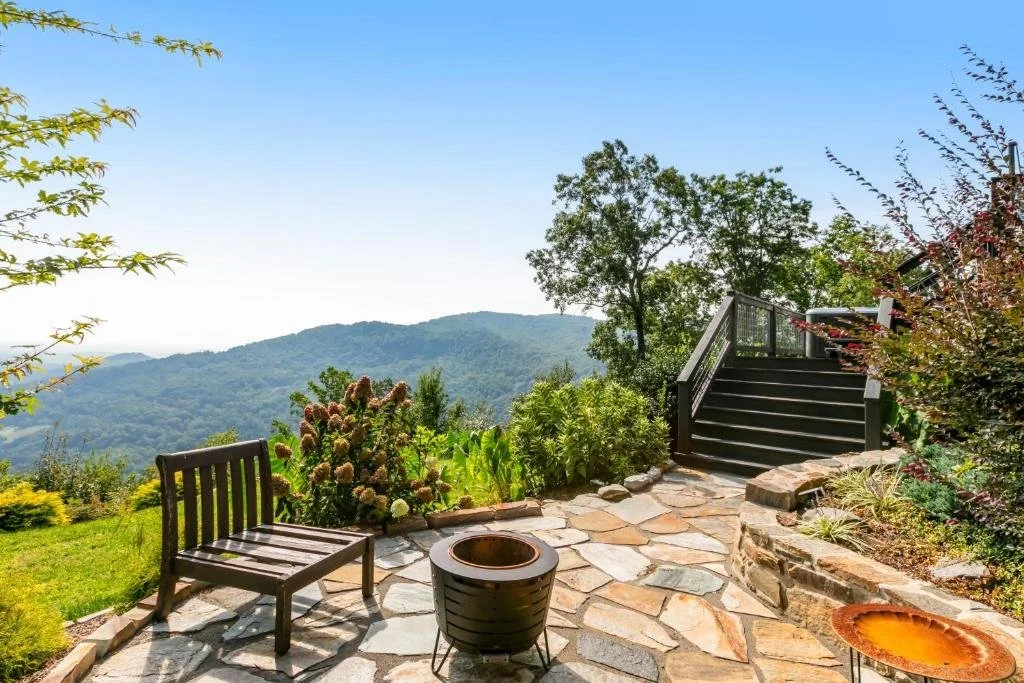 A scenic outdoor patio with a wooden bench, a fire pit, and a stone pathway leading to stairs, overlooking a lush green valley and mountains under a clear blue sky.