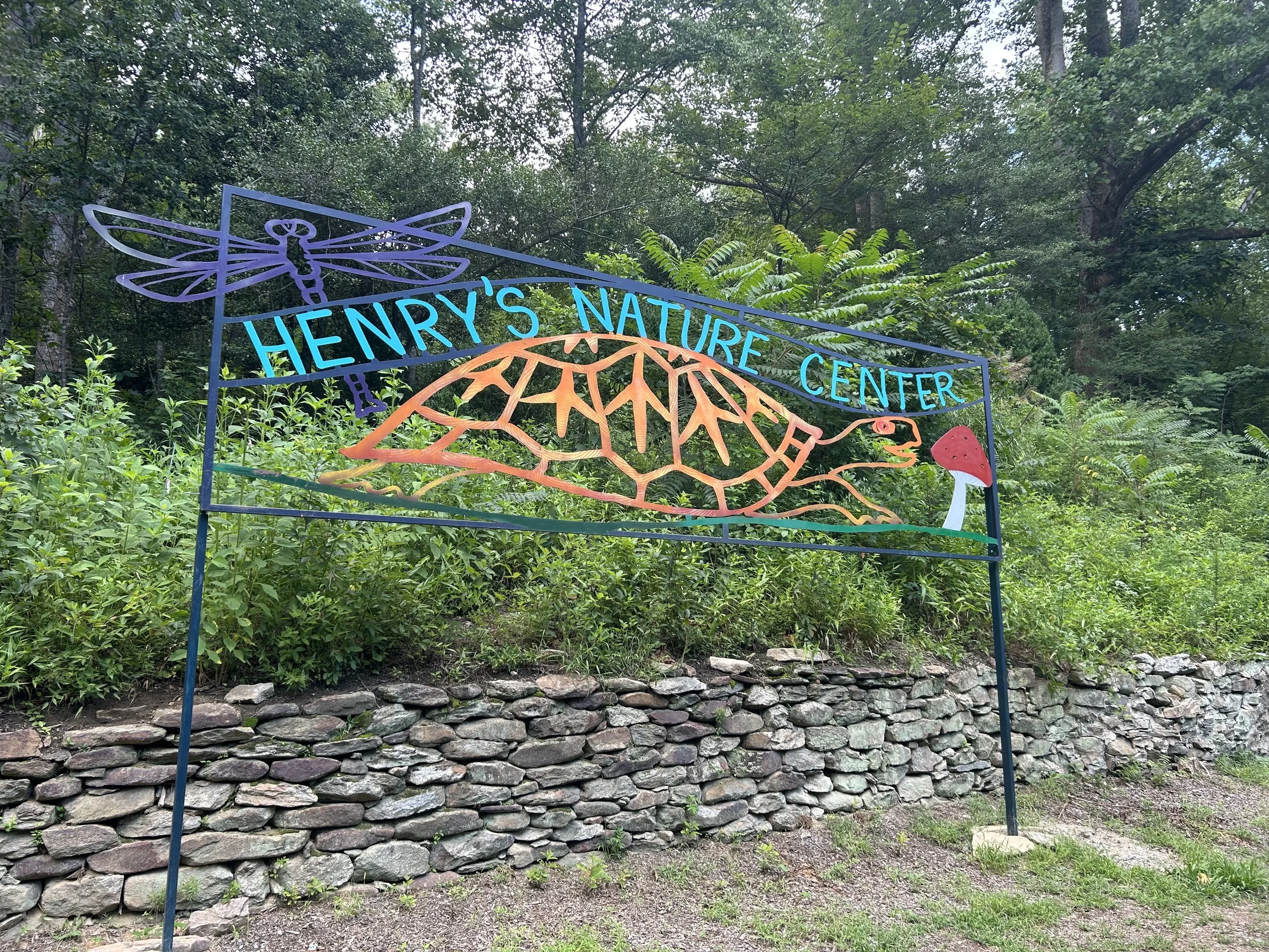 Sign for Henry's Nature Center with a colorful dragonfly, leaf, and mushroom illustrations, set against a background of greenery and trees.