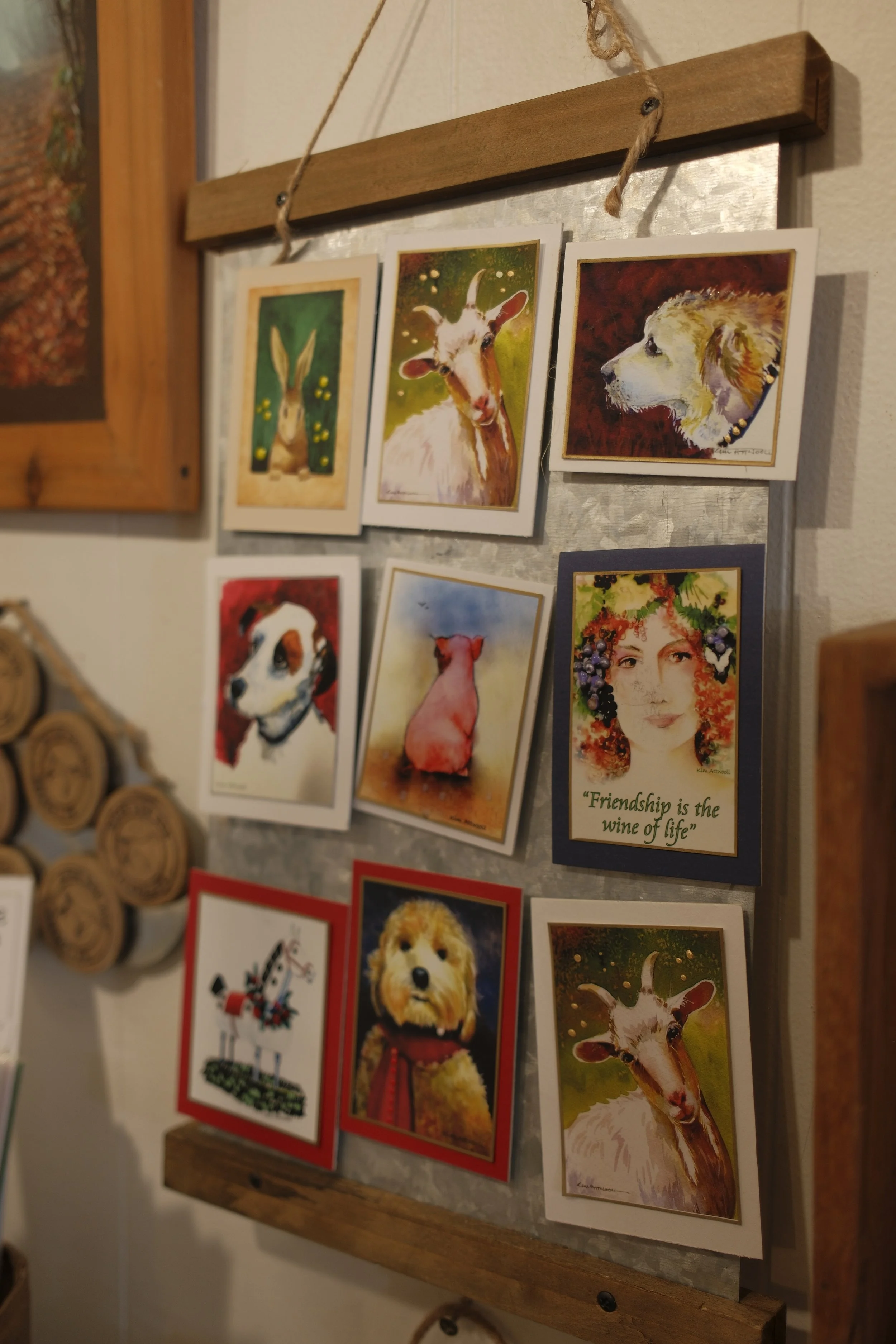 A wooden display frame holding colorful greeting cards with animal illustrations and a quote that says "Friendship is the wine of life". The cards feature a rabbit, goat, dog, cat, and other animals.