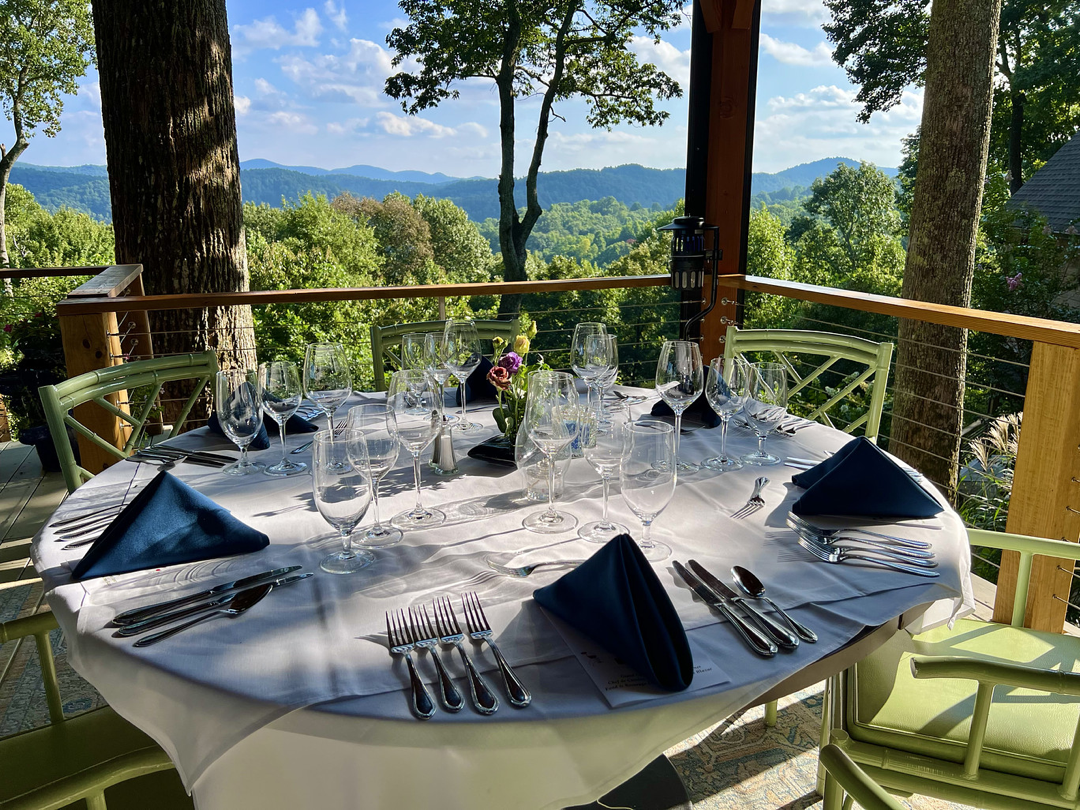 A round table set for a meal with white tablecloth, dark blue napkins, and multiple wine glasses, on a deck overlooking a lush green landscape with mountains in the background.