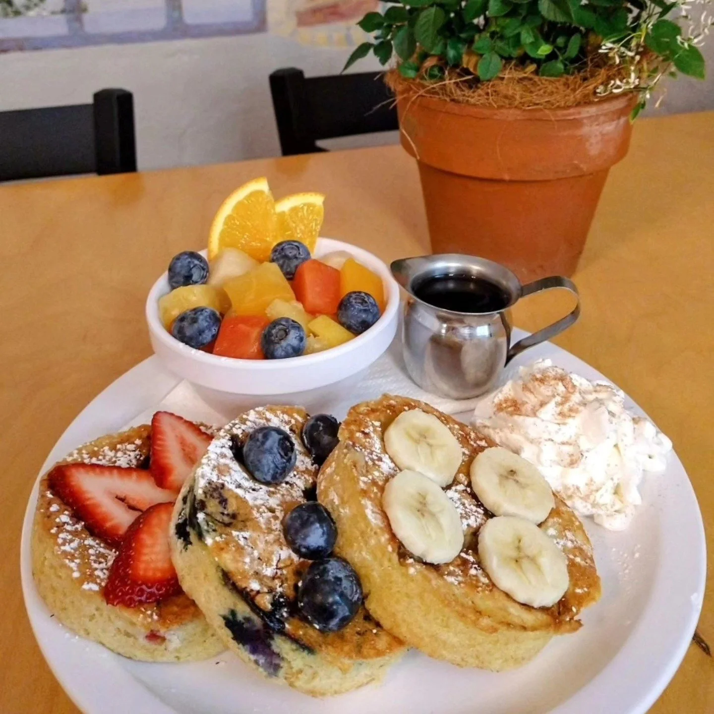 A plate of French toast topped with sliced bananas, blueberries, and powdered sugar, served with a side of whipped cream. A small bowl of fresh fruit salad with orange slices, blueberries, and other mixed fruit, a cup of black coffee, and a small pit