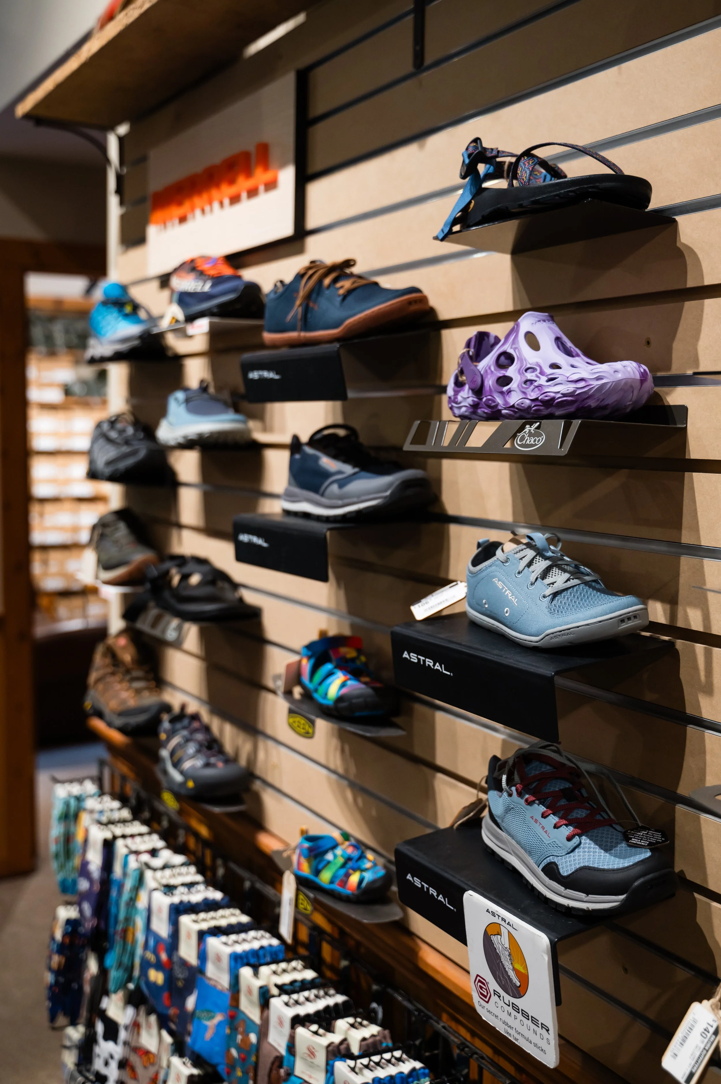 Display of children's shoes, including sneakers and sandals, on black shelves in a retail store. The shelves are mounted on a wooden wall with a pegboard background.