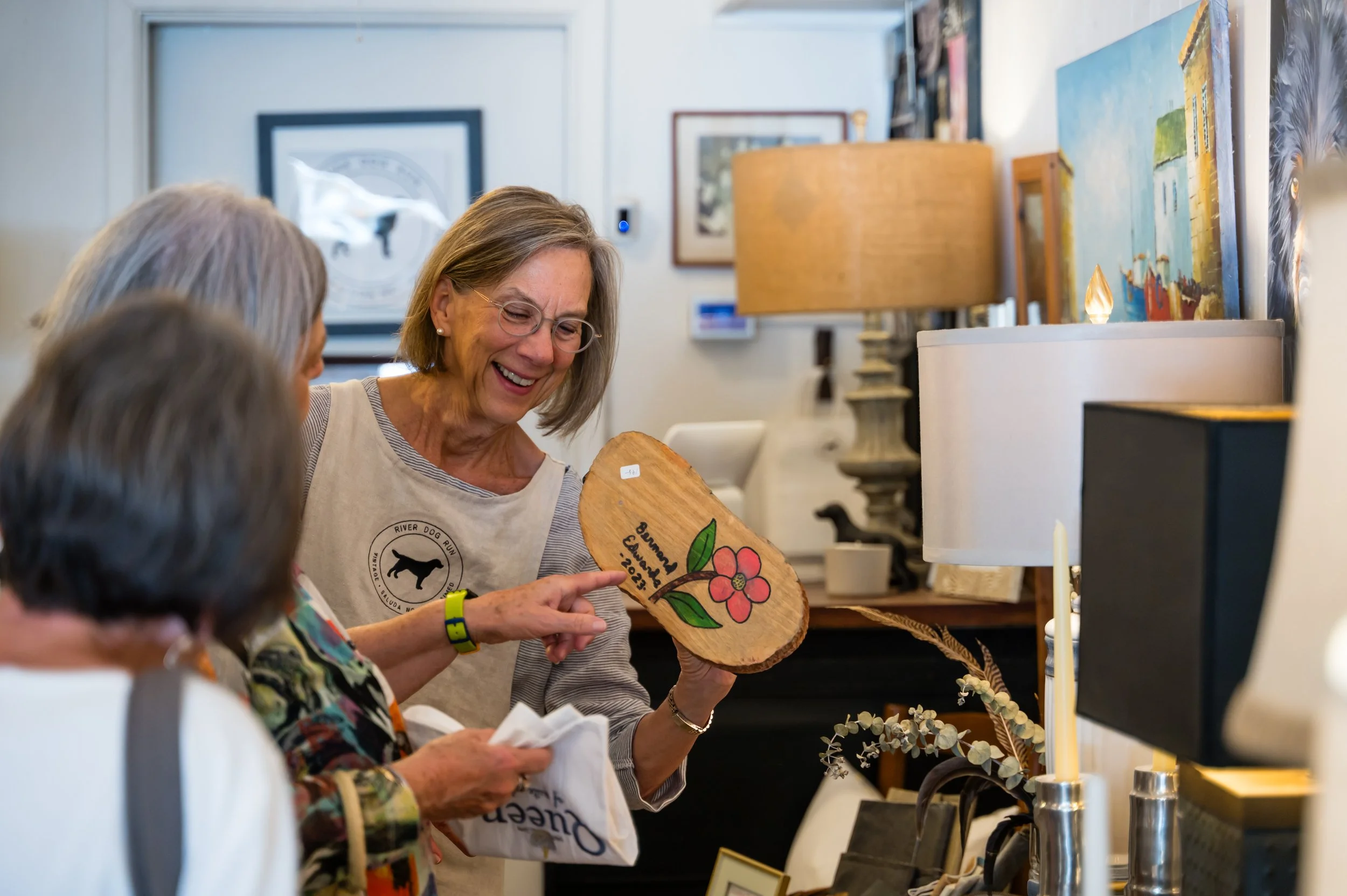 A woman with glasses smiling and holding a wooden plaque decorated with a flower, in an art gallery or studio. Several people are visible, and there are paintings and art supplies in the background.