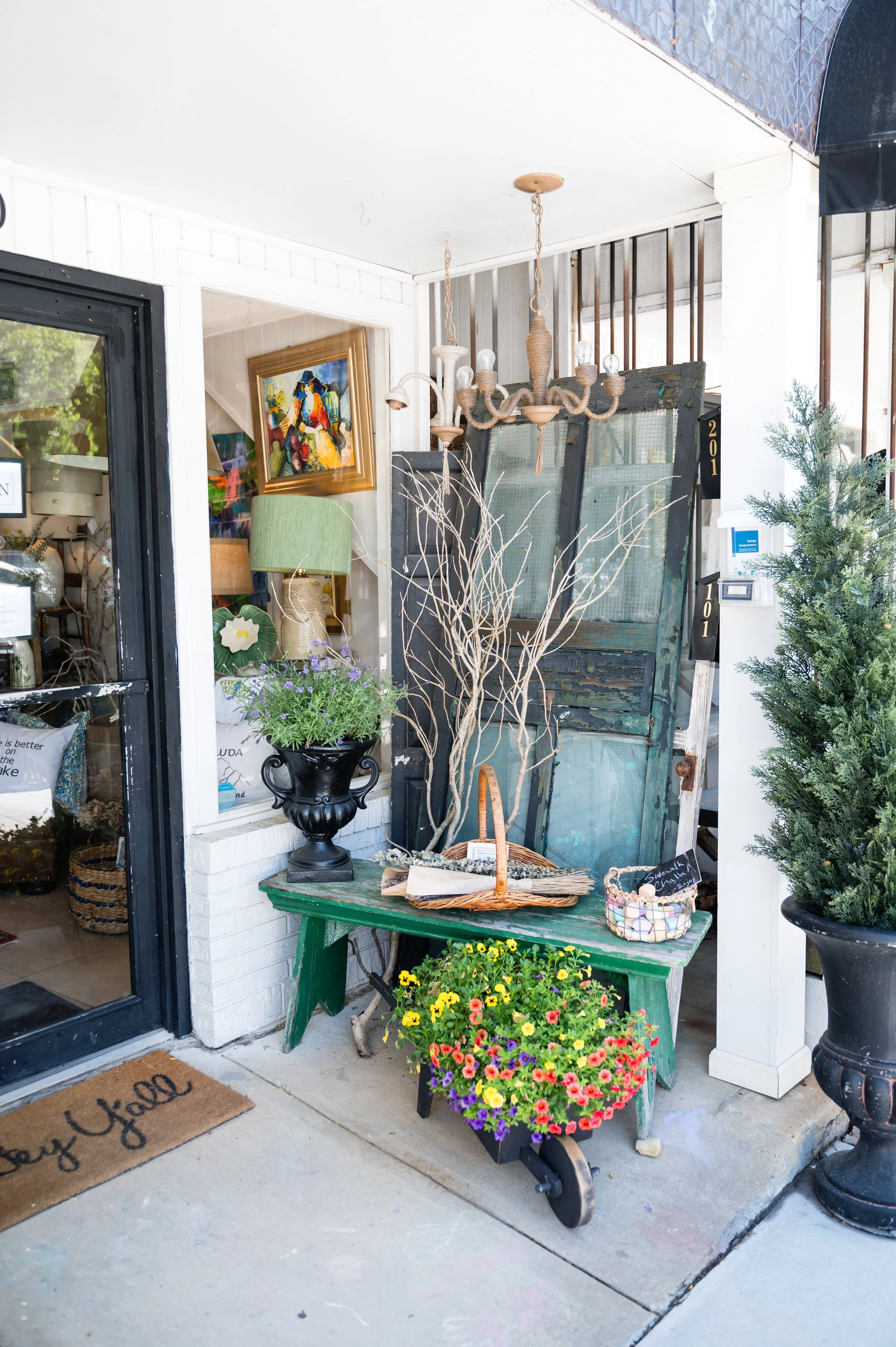 Display of plants and gardening items outside a shop with a green wooden table, a potted flower with colorful blooms, a black and white painted basket, a dried tree branch, and a wicker basket with stones inside.