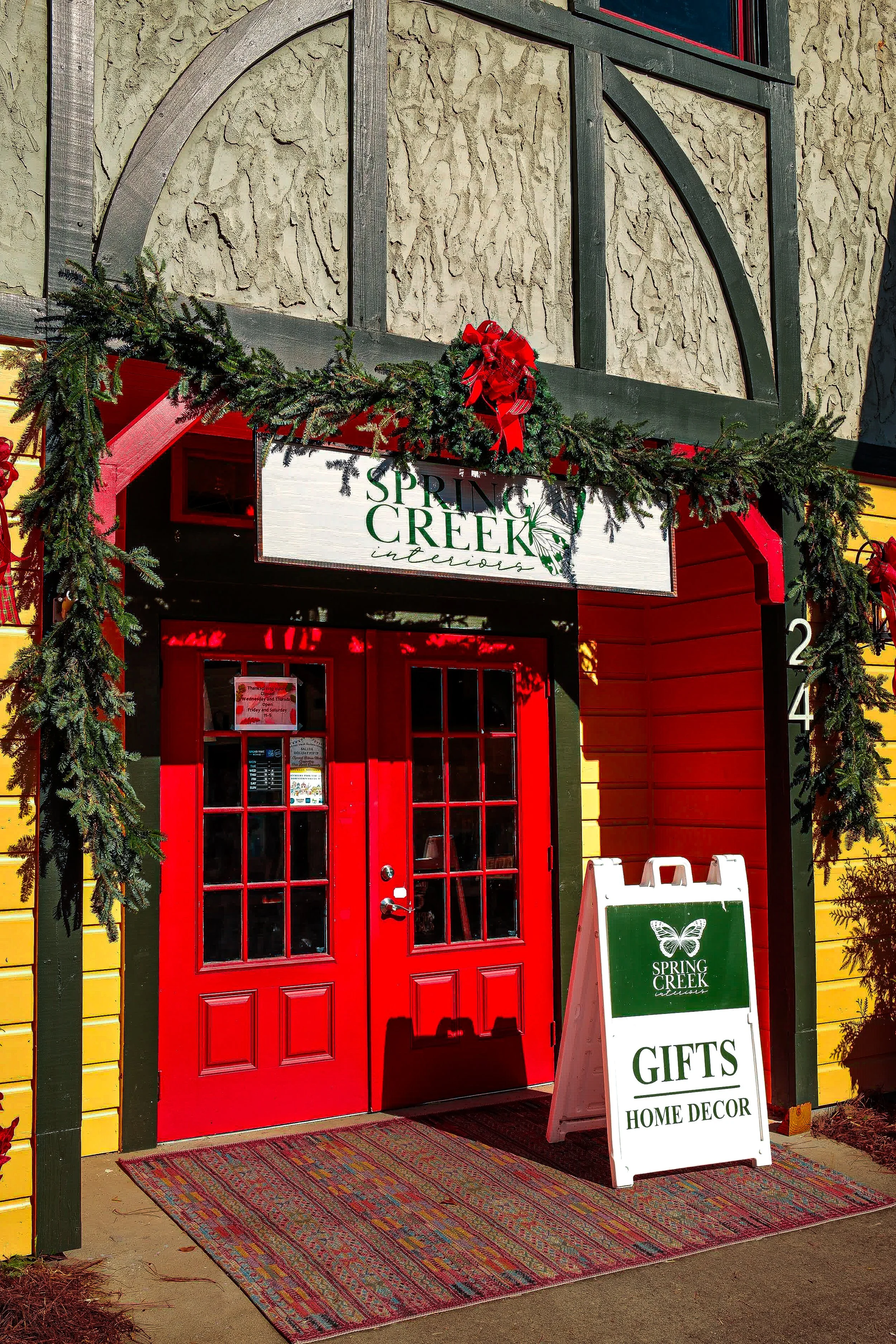 Red door with multiple glass panes at the entrance of a shop called 'Spring Creek Interiors' decorated with greenery and a red bow as part of Christmas holiday decorations. There is a white sandwich board sign nearby advertising gifts and home decor.