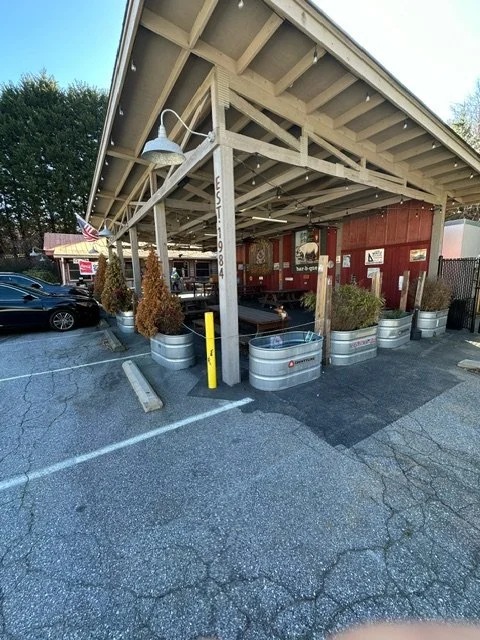 Parking lot with a wooden covered outdoor seating area, planters made of metal, and a red building behind with various signs.