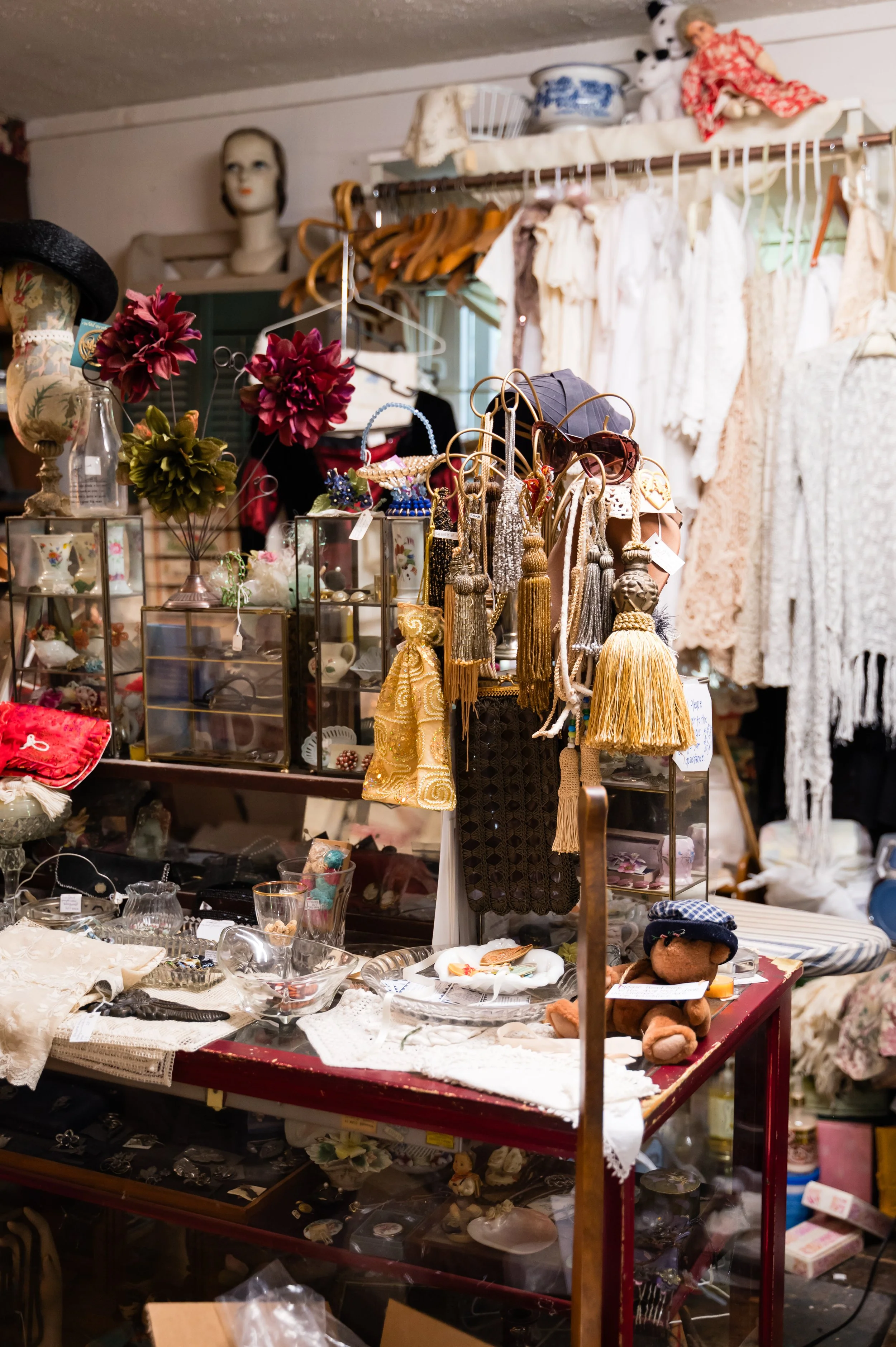 A vintage thrift store display with various secondhand items including jewelry, glasses, dolls, clothing, and decorative objects on shelves and tables.