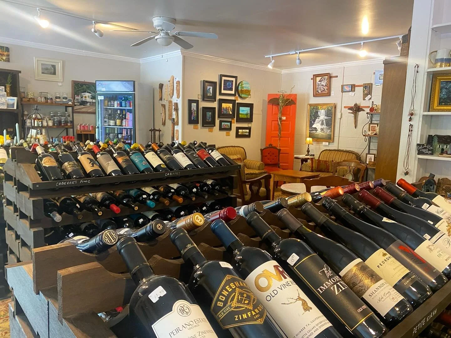 Interior of a wine shop with shelves displaying multiple bottles of wine, framed artwork on the walls, and a seating area with vintage-style couches and a coffee table.