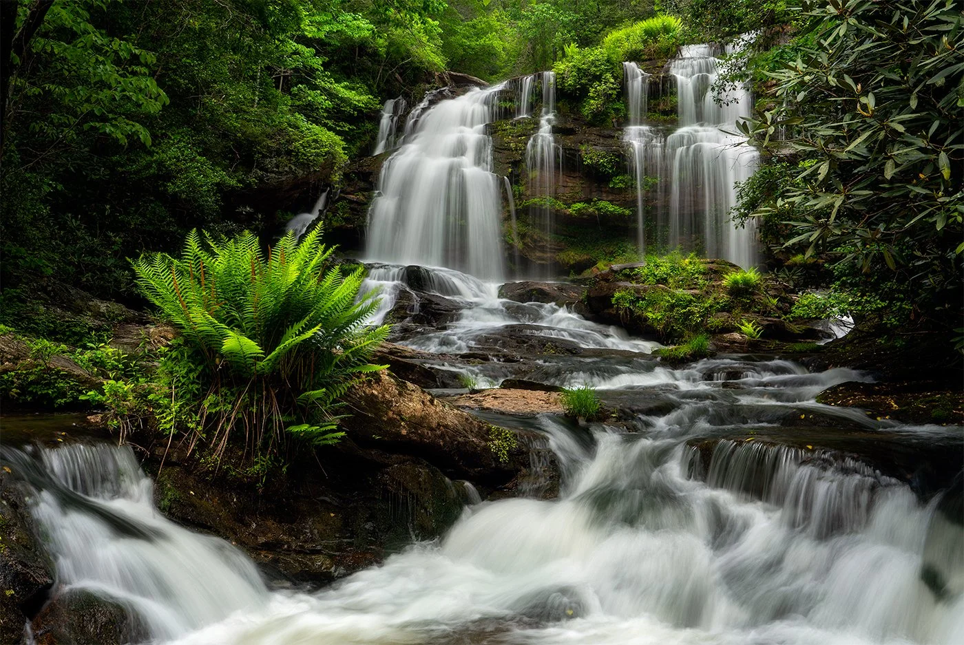 A scenic view of a multi-tiered waterfall flowing through a lush green forest with rocks and vibrant foliage.
