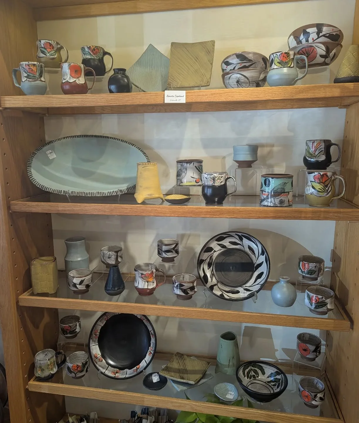 Display of various ceramic and glass dishware on wooden shelves, including mugs, bowls, plates, and cups with floral and abstract patterns.