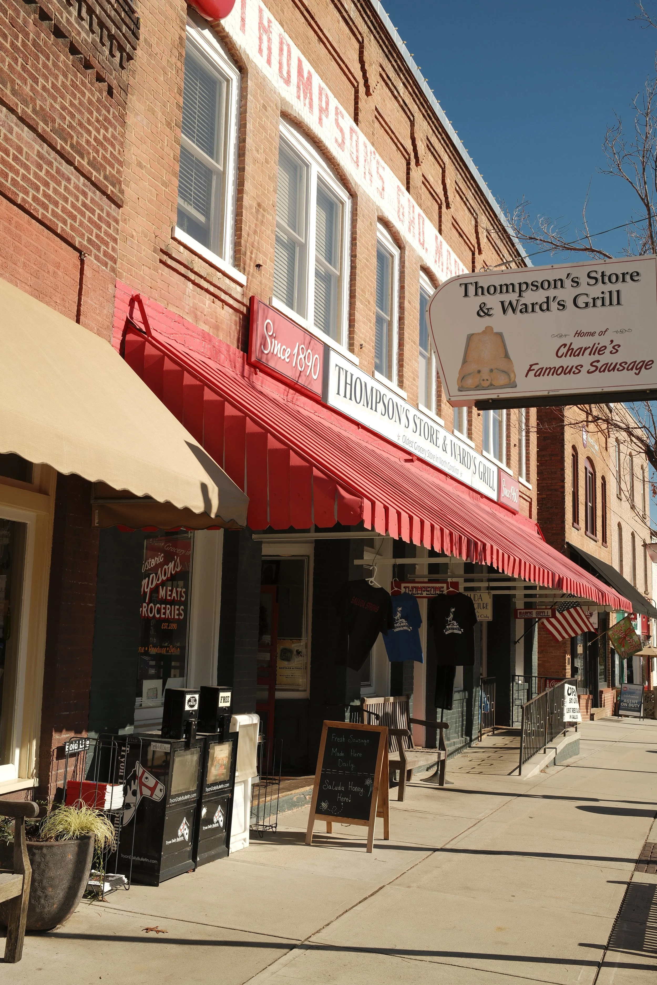 Street view of a brick building with storefronts, red awnings, signs indicating a store established in 1890, and an outdoor display with t-shirts and a chalkboard sign in front of the store. The sky is blue and there are a few trees with no leaves.