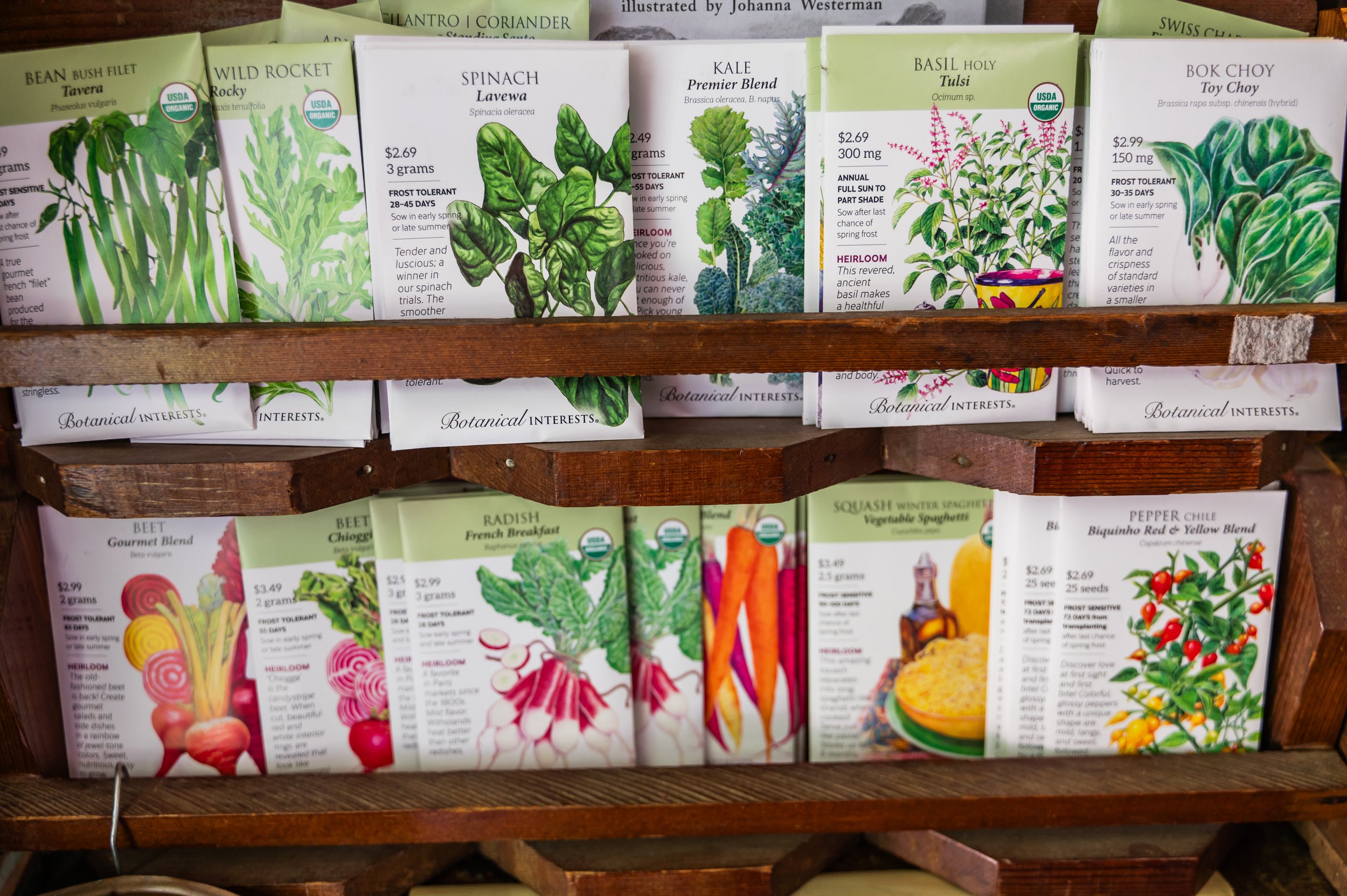 A wooden shelf holding multiple packets of organic seed varieties, including spinach, kale, basil, bok choy, and rocket. Each packet features colorful illustrations of the vegetables and information about their growing conditions.