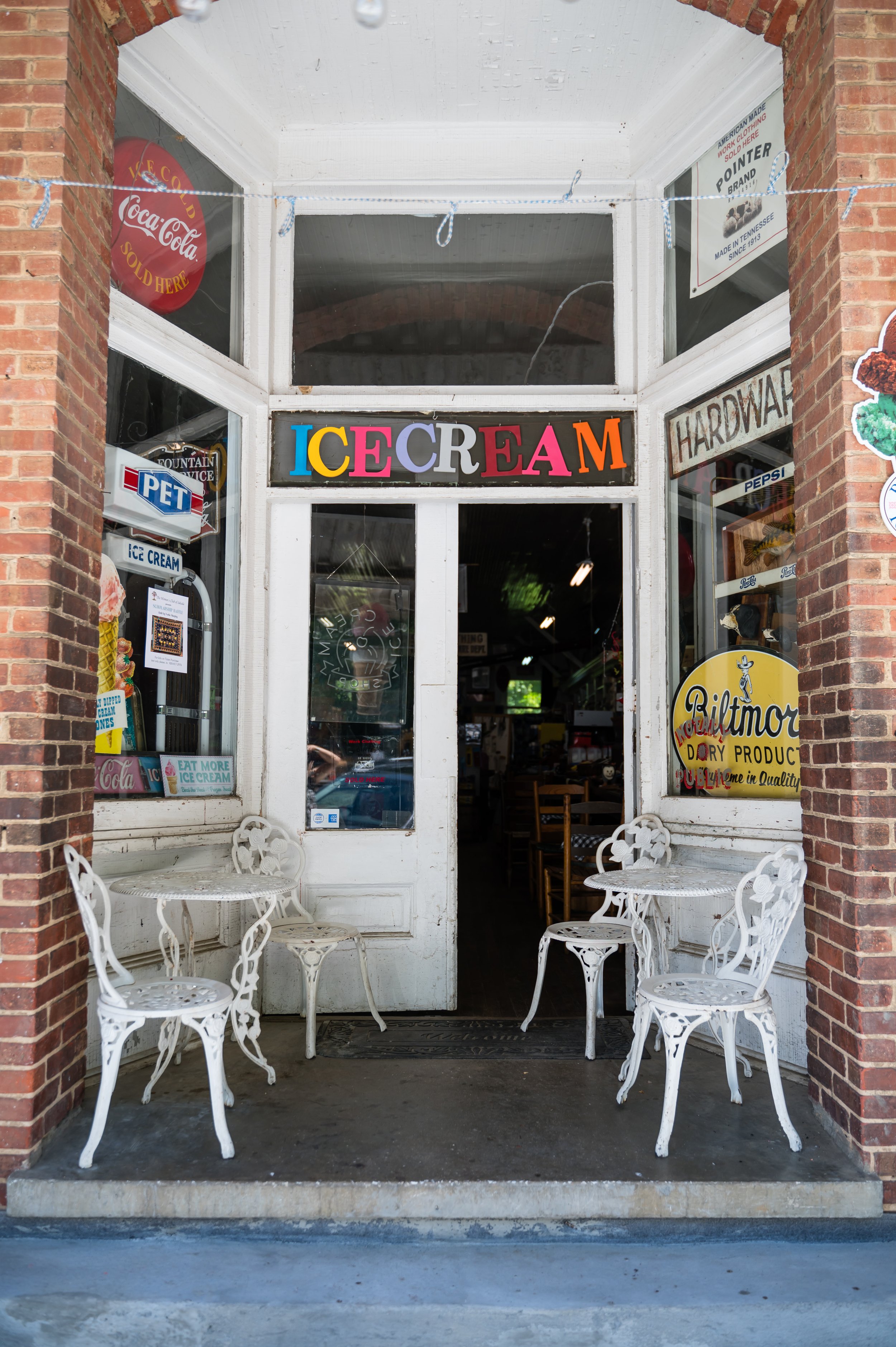 The storefront of an ice cream shop with a white door and windows, decorated with colorful signs and vintage decor, with two white metal chairs and tables outside.