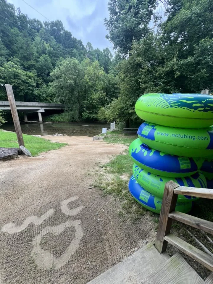 Stack of inflatable tubes beside a dirt path leading to a river, with a bridge and dense green trees in the background.