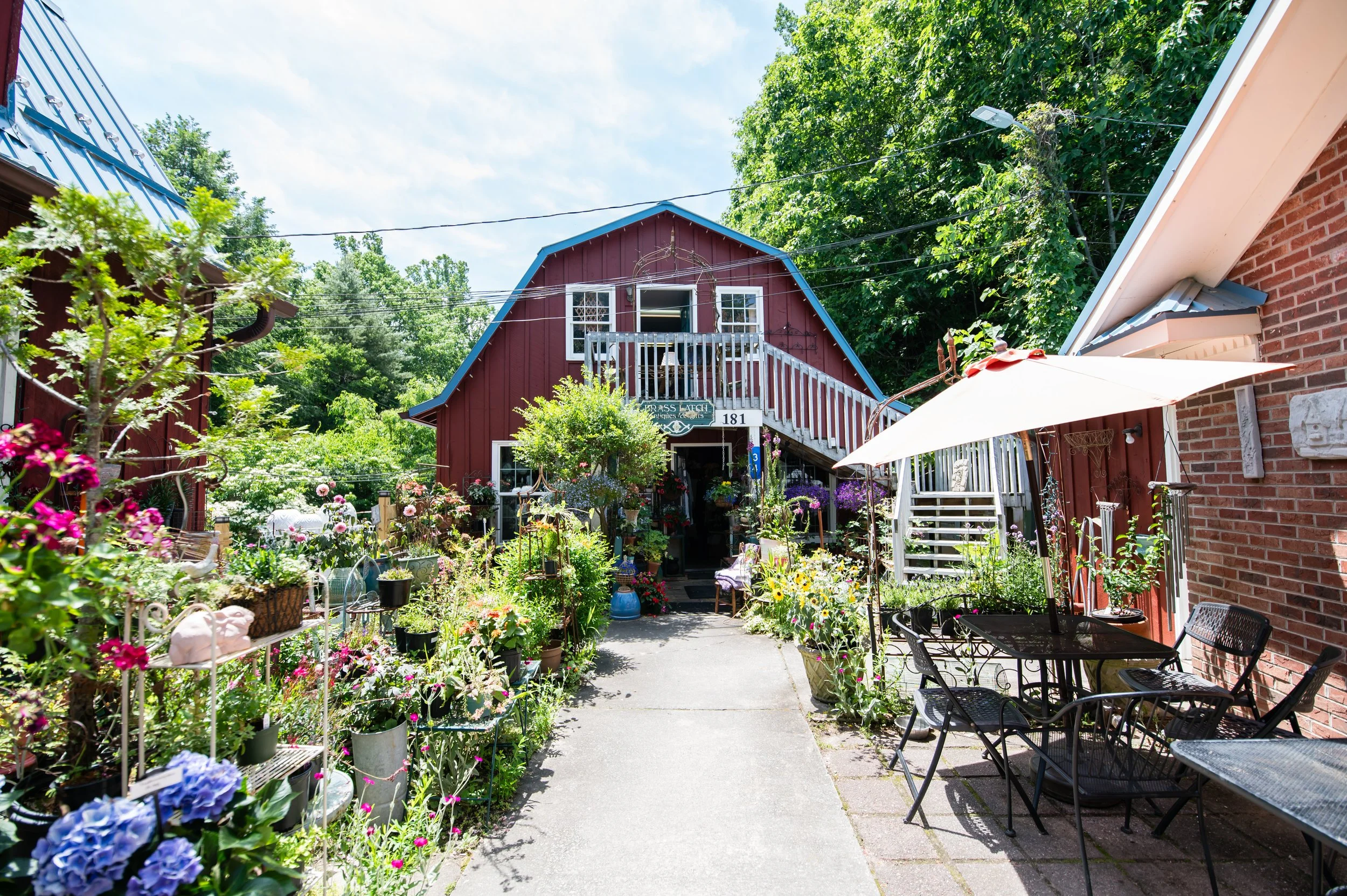 A backyard garden with a flower bed, patio furniture, and a red barn-style building in the background, surrounded by lush green trees and plants.