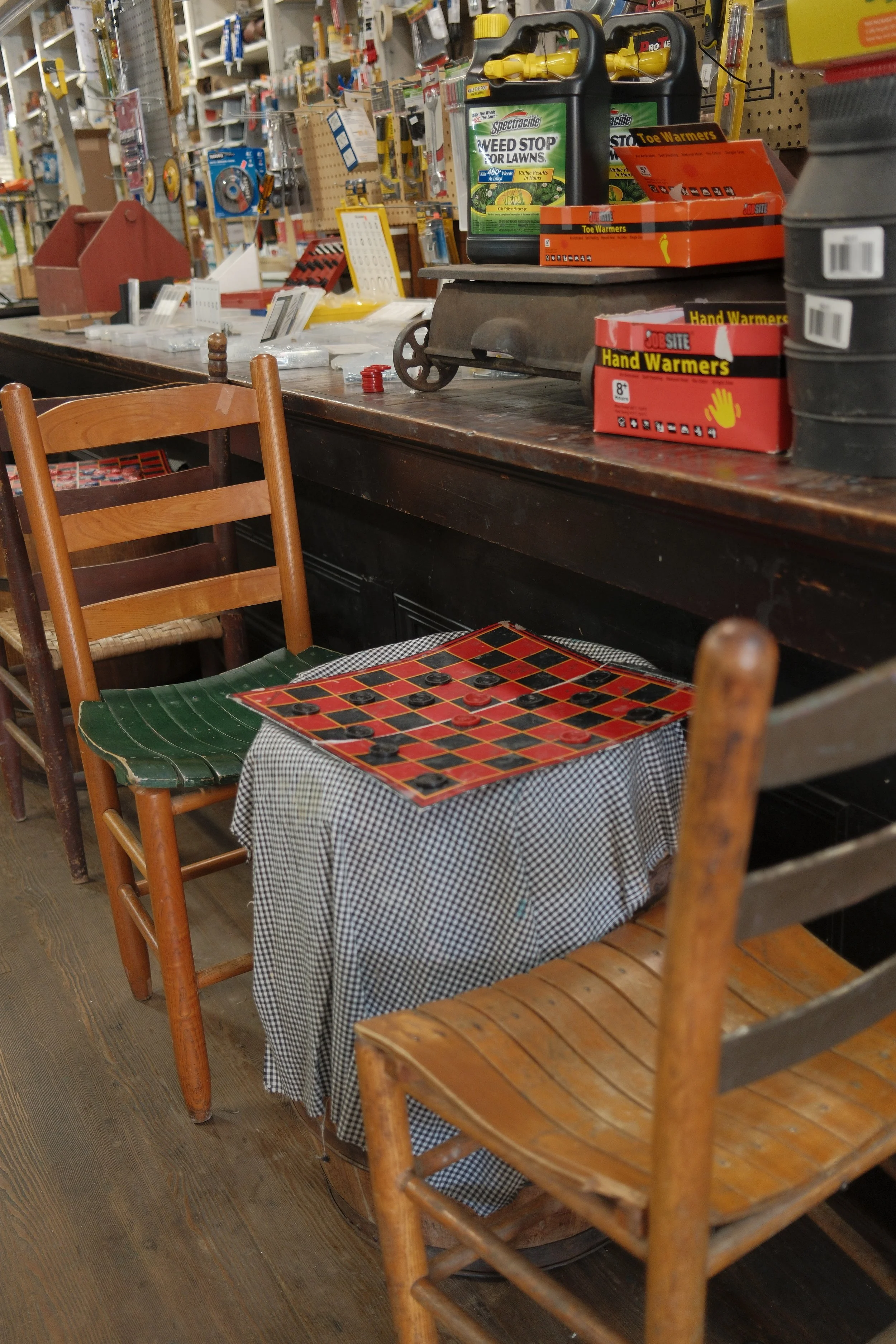A cluttered wooden table in a hardware store with various garden supplies and hand warmers, surrounded by wooden chairs on a wooden floor.