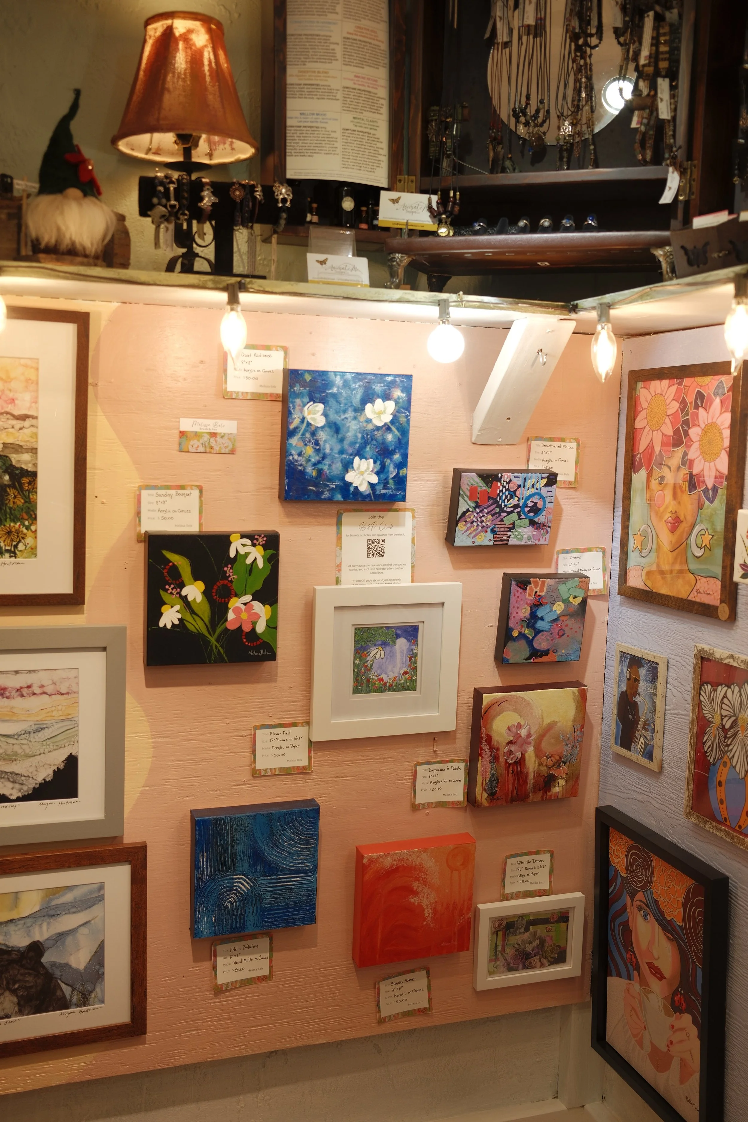 An art gallery display wall with various small paintings and artwork, some framed and some unframed, with prices and labels. Some paintings depict flowers, abstract patterns, and landscapes.