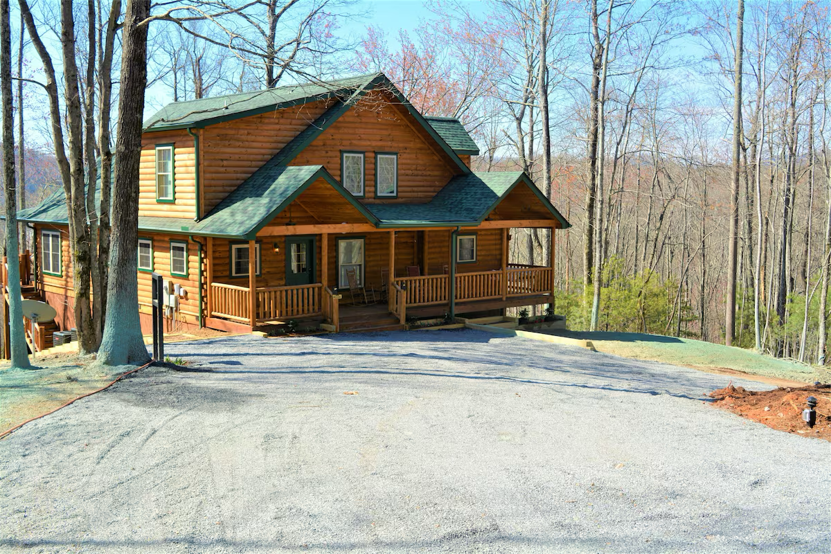 A two-story wooden house with a front porch, surrounded by leafless trees in a wooded area during winter or early spring.