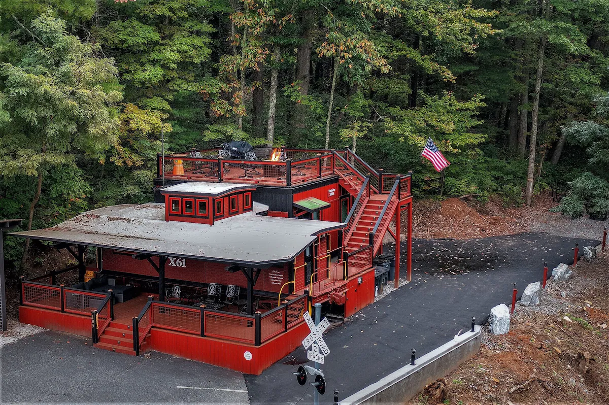 A small vintage train station with a red and black exterior, located next to a wooded area, featuring an American flag, a covered waiting area, and a railroad crossing sign, with a short train platform and stairs leading to an upper deck.