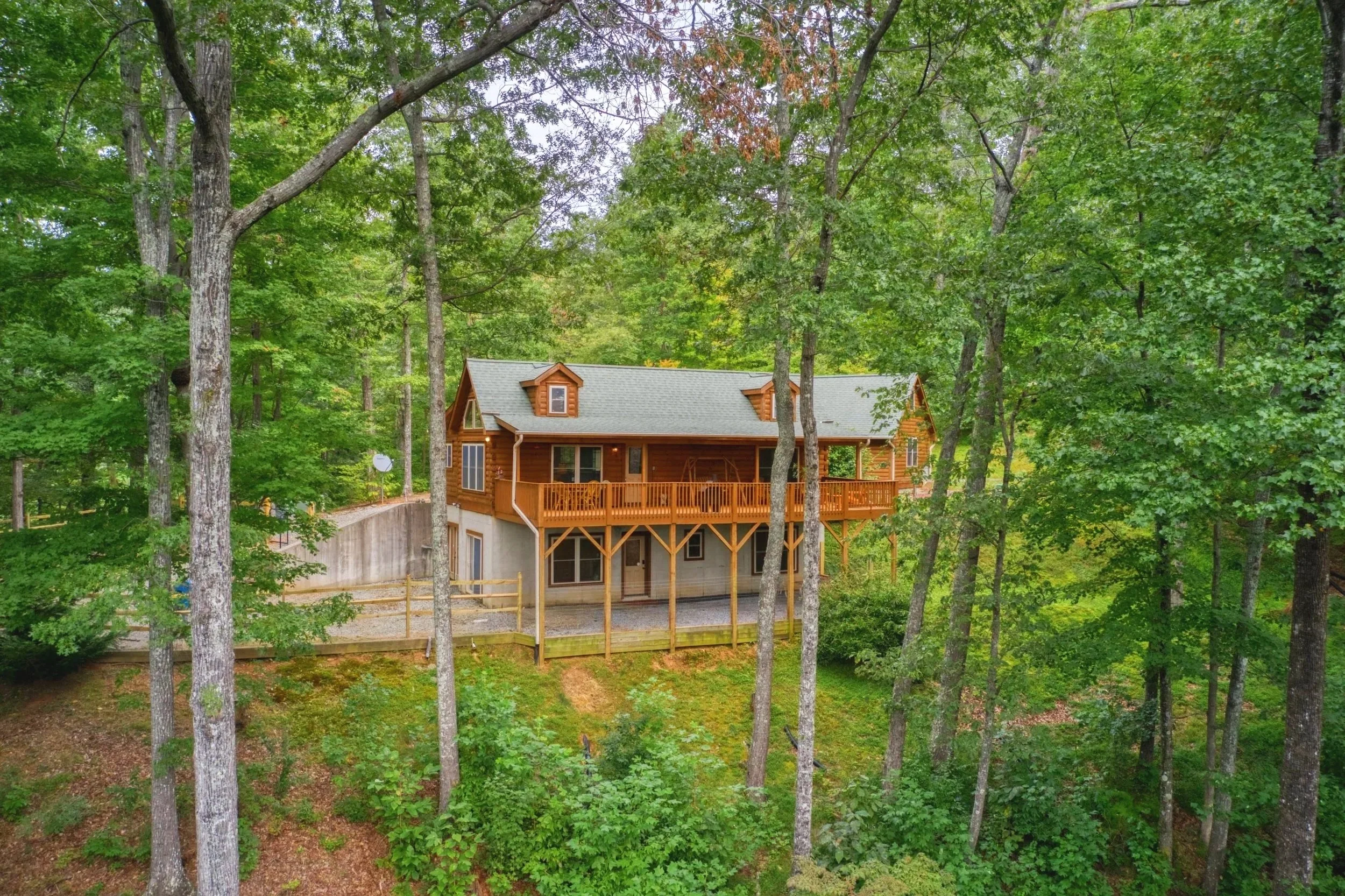 A wooden house with a loft and a deck is surrounded by tall trees and dense green foliage in a forested area.