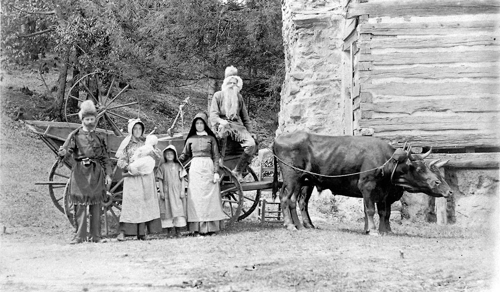 A black-and-white photo of a family with four children and one adult, standing in front of a wooden barn. The adult man with a beard is sitting on a cart pulled by two oxen. The children are dressed in old-fashioned clothing, with the girls wearing long dresses and bonnets, and the boy wearing a jacket and hat.