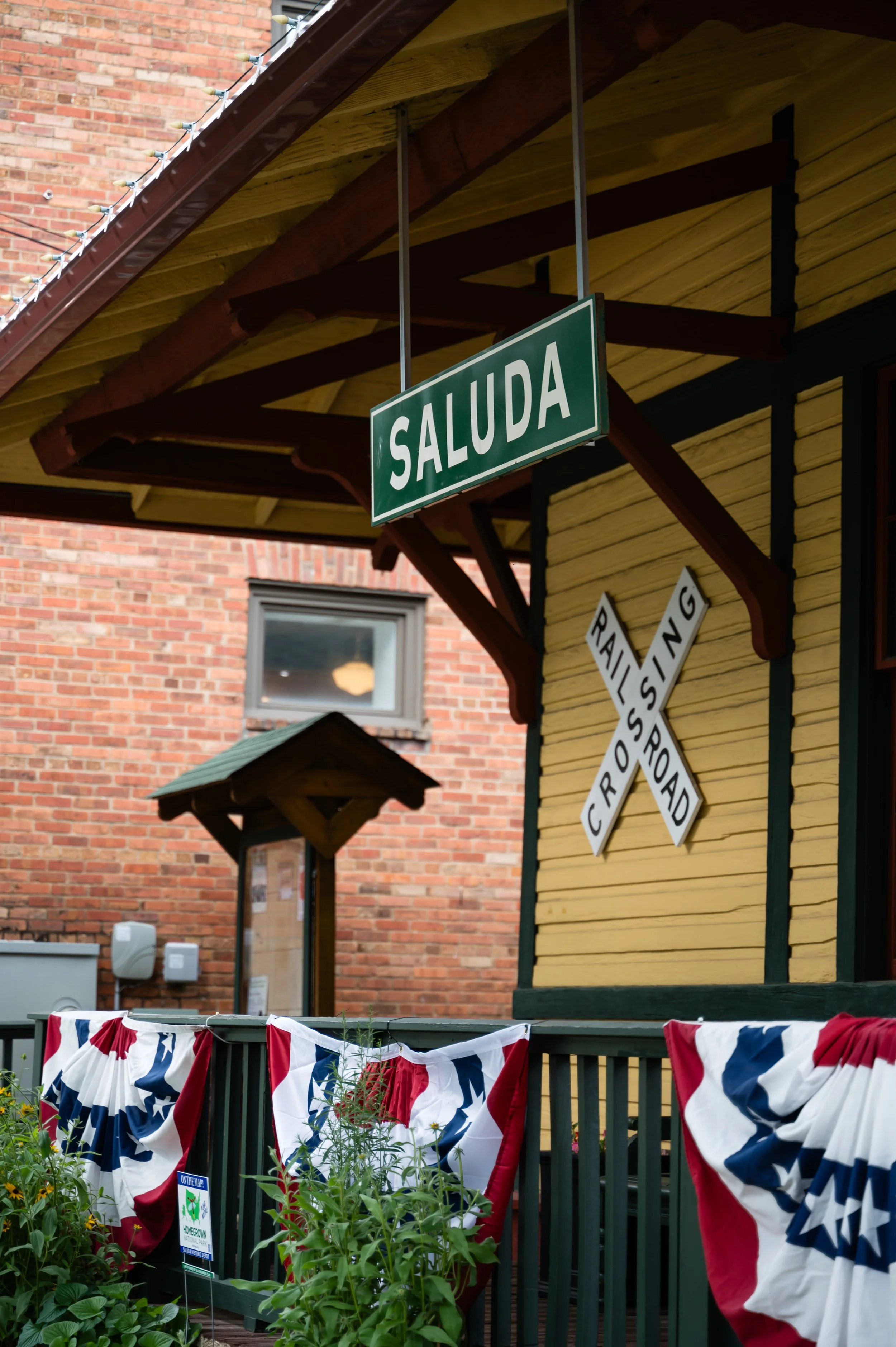 A green sign with white letters saying 'SALUDA' hanging under the roof of a yellow building with dark trim. Below, red, white, and blue patriotic bunting decorates the black railing of a porch or balcony in front of a garden with various plants. A brick building with a window and some utility boxes is visible in the background.