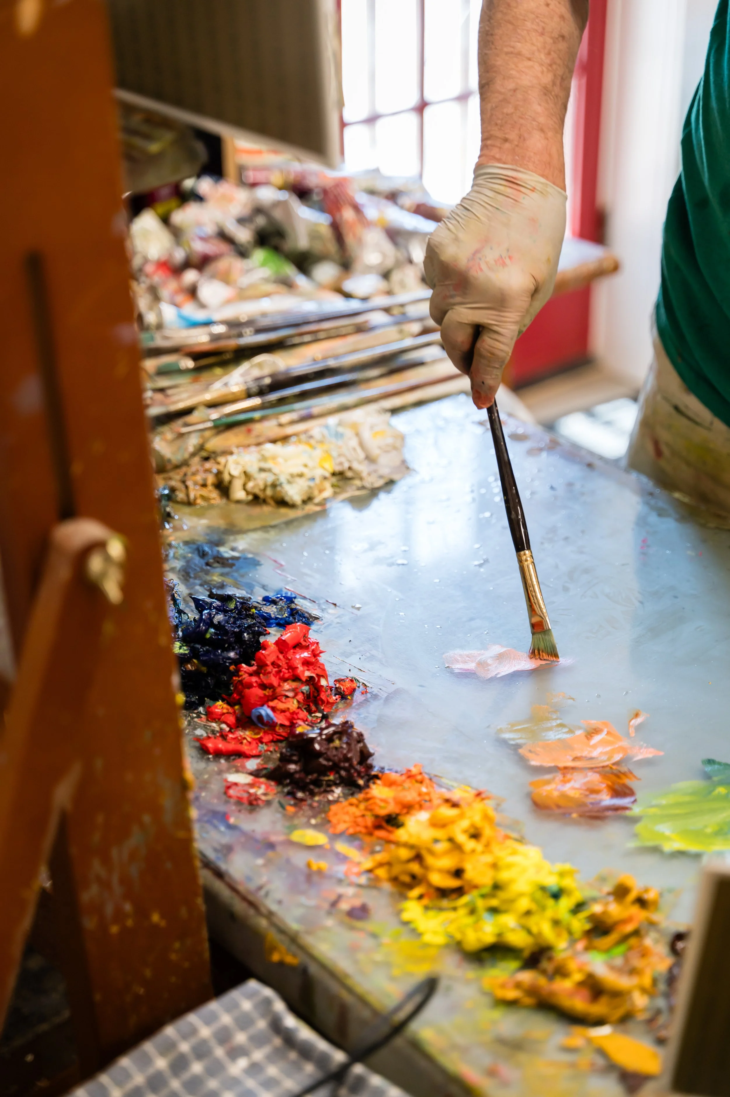 An artist's hand holding a paintbrush, creating a colorful painting on a large canvas or palette with various shades of paint, in a bright indoor studio.