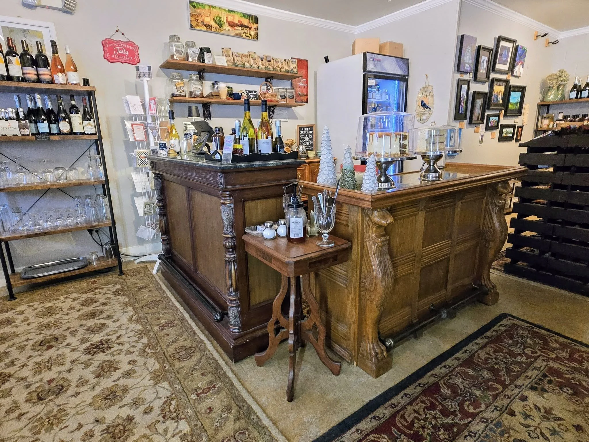 Interior of a small cafe or bakery with a wooden counter, shelves stocked with wine bottles and baked goods, and various framed pictures on the walls. There are also decorative Christmas trees on the counter.