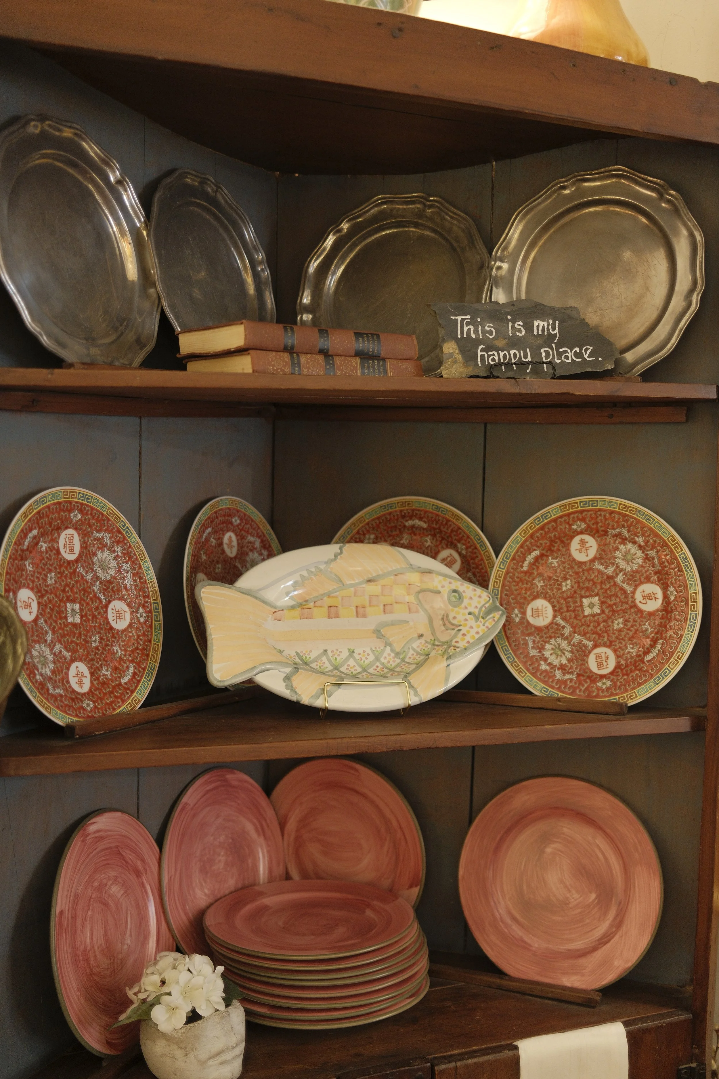 A wooden bookshelf displaying various decorative plates, vintage books, a fish-shaped dish, and a small flower arrangement, with a sign that reads 'This is my happy place'.