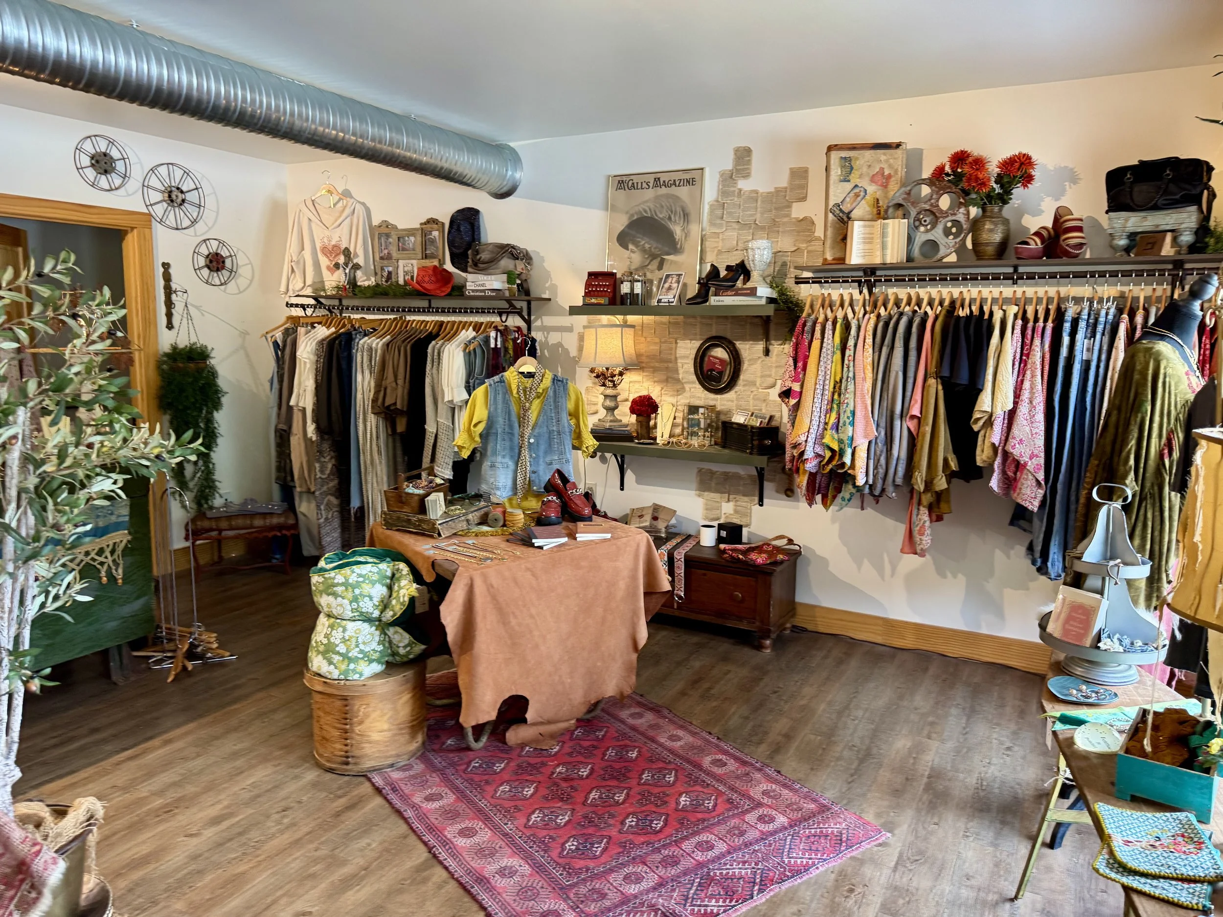A cozy boutique store with clothing racks, decorative wall art, and a table displaying accessories, shoes, and scarves. The store has wooden floors, a red patterned rug, and vintage decor.