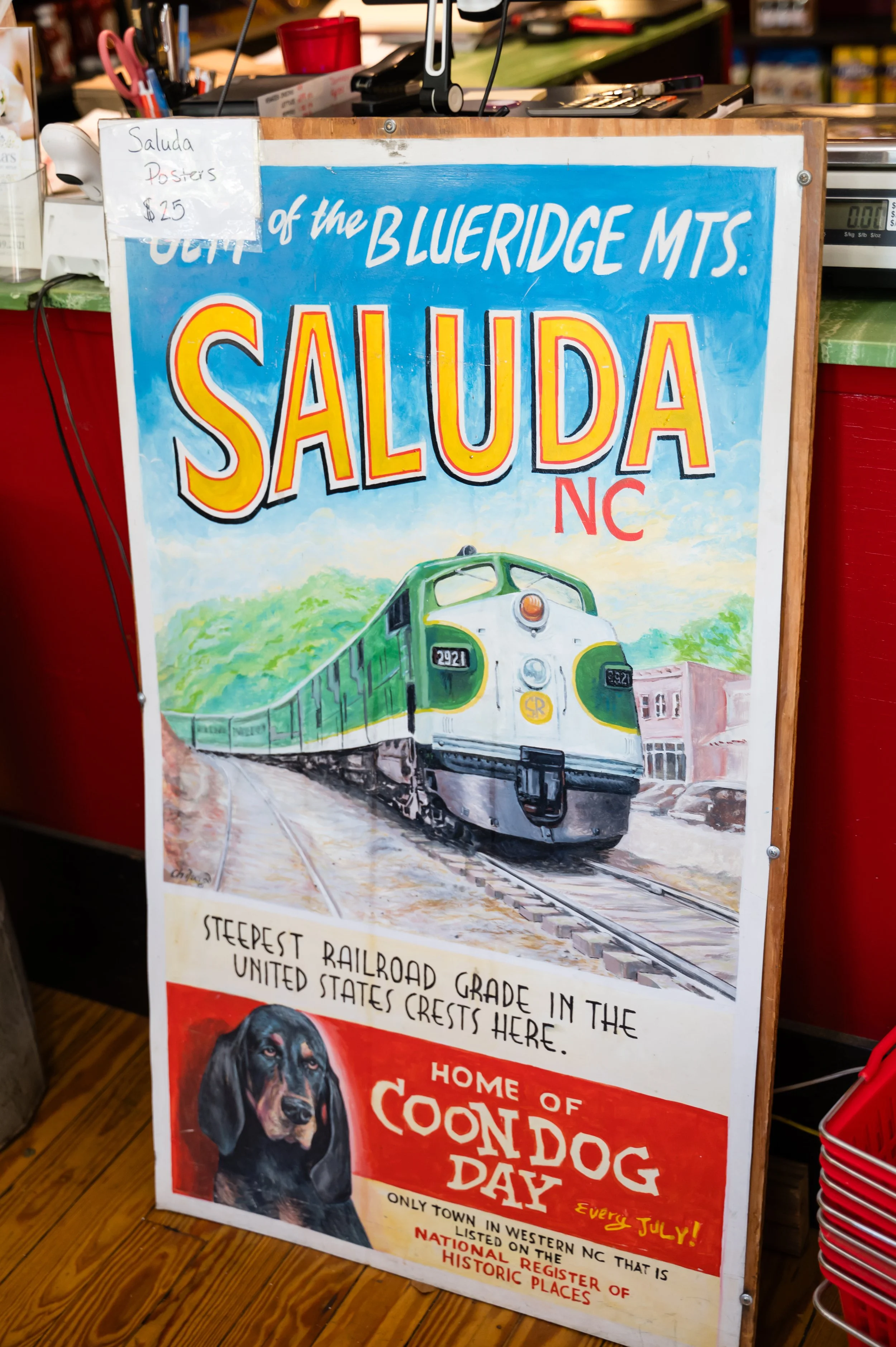 A colorful sign promoting Saluda, North Carolina. It features a train illustration and mentions the town's historic significance, including its steep railroad grade and status as a national register of historic places.