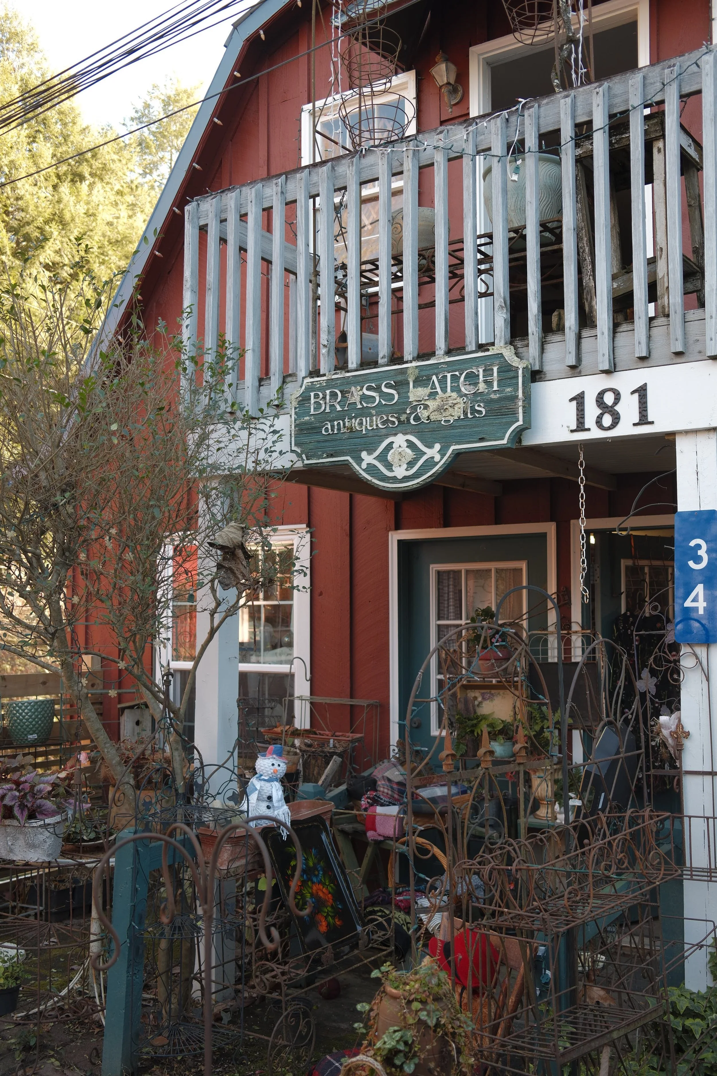 A house with red painted wooden exterior and white window frames. A small balcony with a rustic wooden railing displays various antique metal and ceramic items. Decorations and garden objects are scattered in the front yard, including a snowman figure, metal chairs, and potted plants. A green vintage sign reads 'Brass Latch antiques & gifts'. The house number '181' is visible, and there are trees and electrical wires in the background.