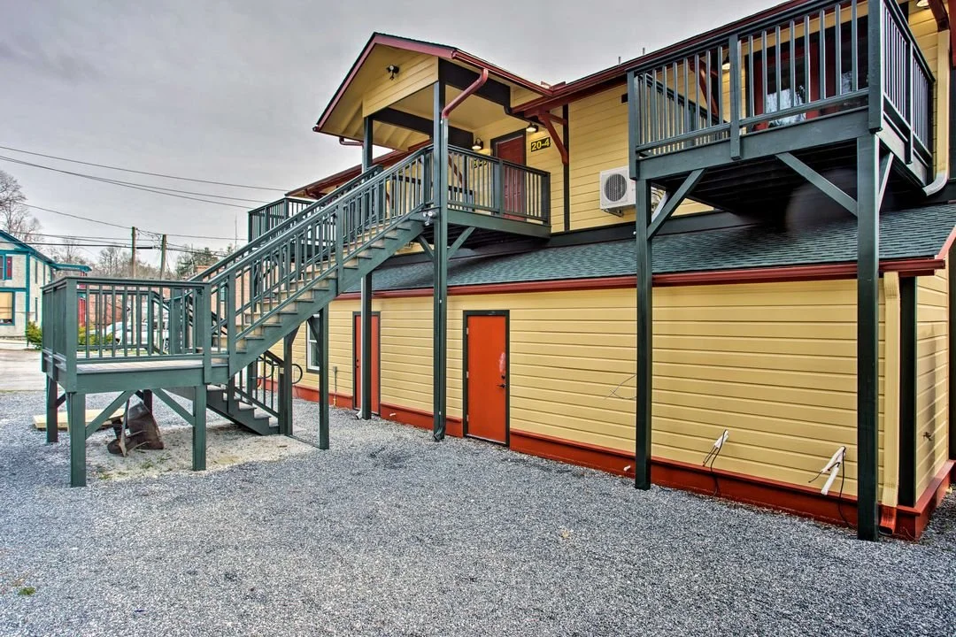 Yellow building with green railings and stairs, and orange door, gravel ground, cloudy sky.