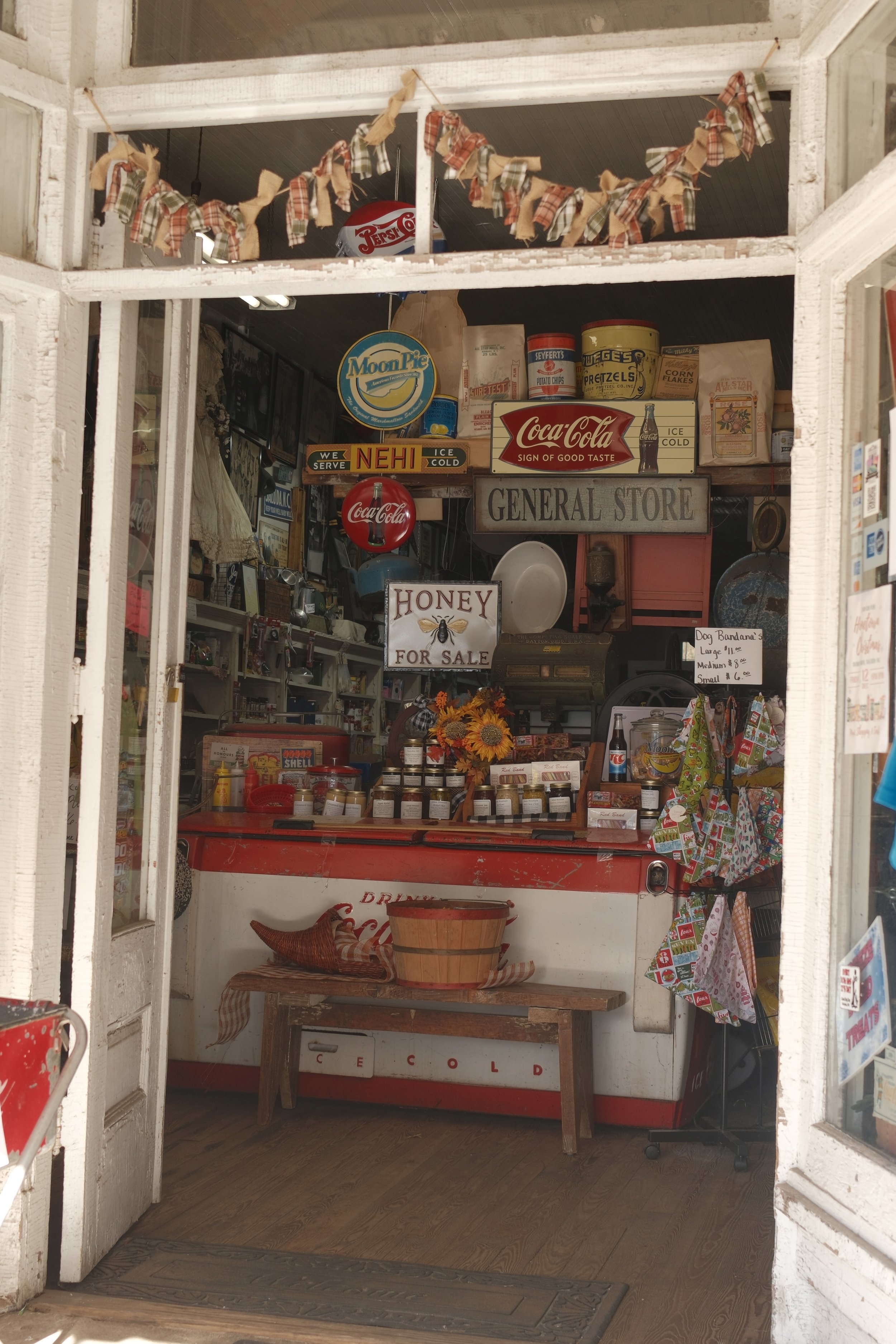 View from outside a small general store with opening double doors. Inside, vintage Coca-Cola signs, nostalgic items, and a display of honey jars are visible. The store has a rustic, nostalgic feel with wooden floors and shelves.