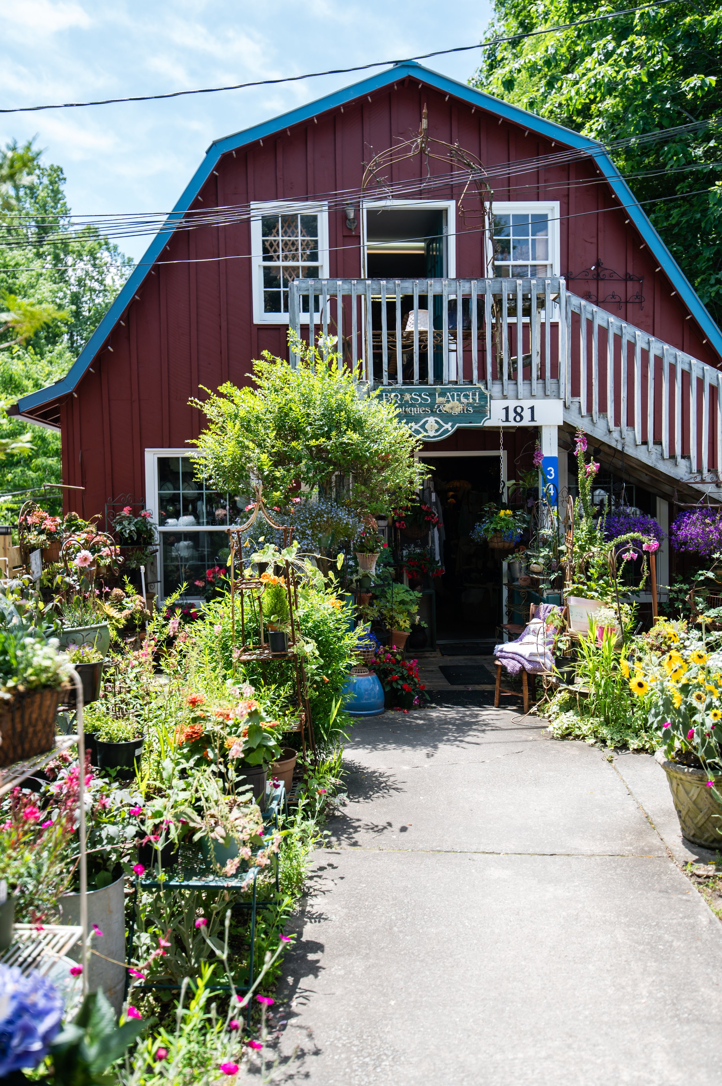 A red two-story building with blue roof trim and white window frames, surrounded by a garden with colorful flowers and plants, with an open door on the second floor and a sign that reads 'Brass Latch Antiques & Gifts' at the front.