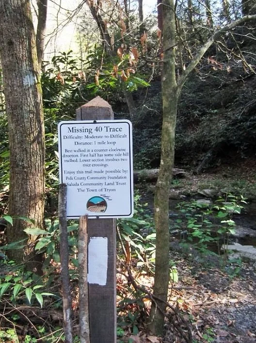 A trail sign reading "Missing 40 Trace," indicating a moderate to difficult trail, one mile long, passing through a wooded area near a creek and surrounded by trees and bushes.