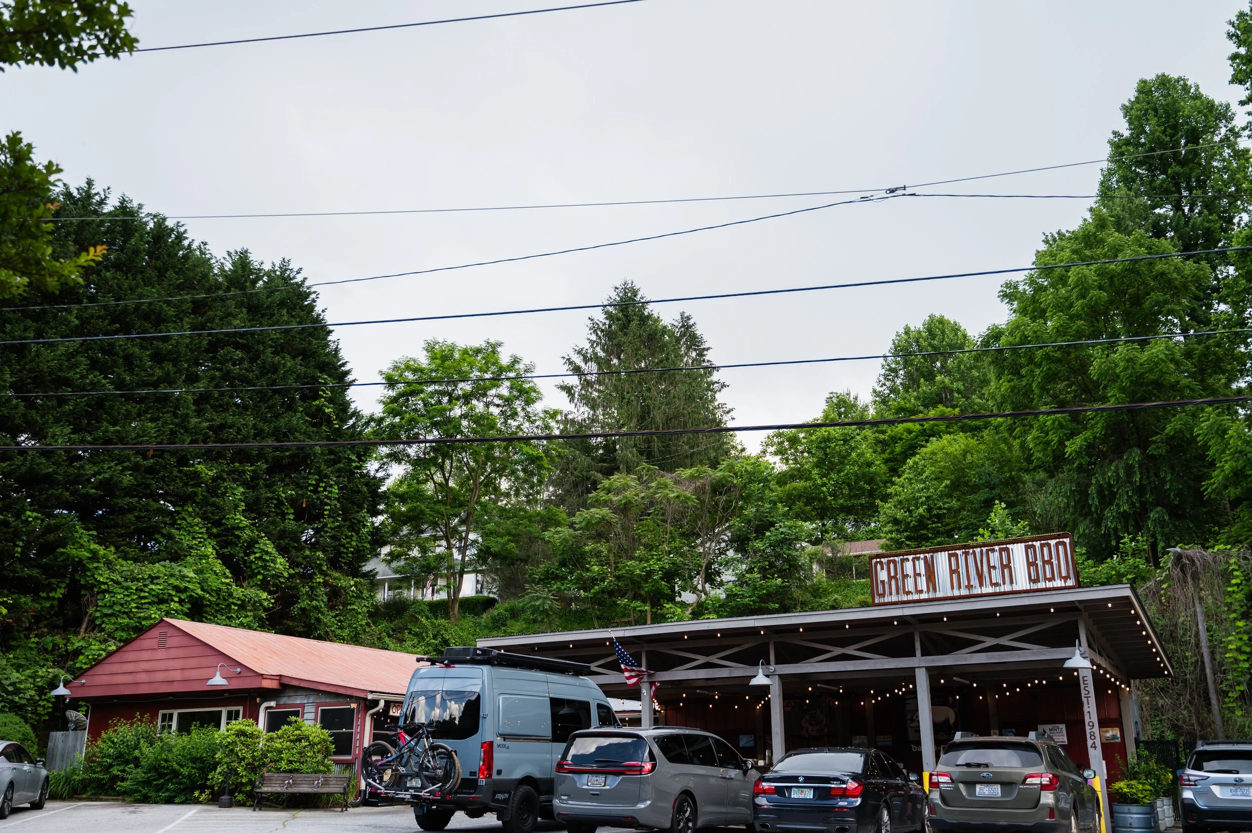 Parking lot in front of a restaurant named Green River BBQ, with several cars and a van with bicycles mounted on the back. Behind the restaurant, there are lush green trees and a cloudy sky.