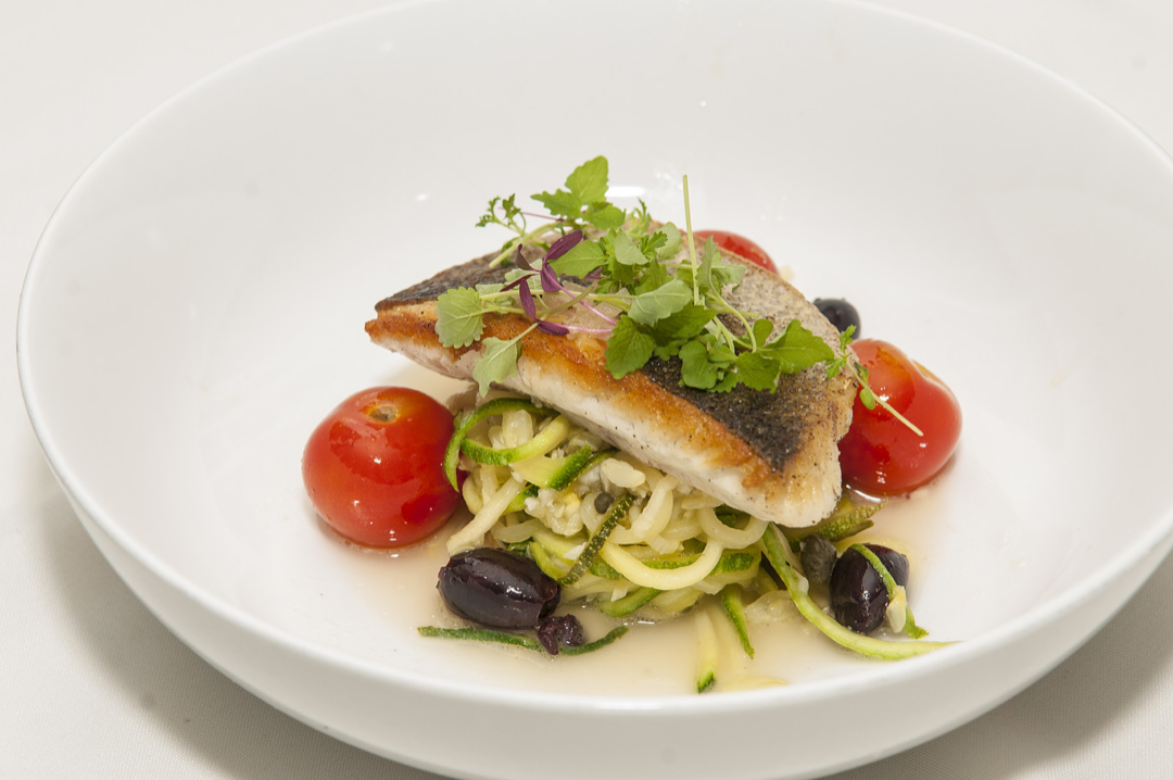Pan-seared fish fillet on zucchini noodles, cherry tomatoes, black olives, garnished with microgreens in a white bowl.