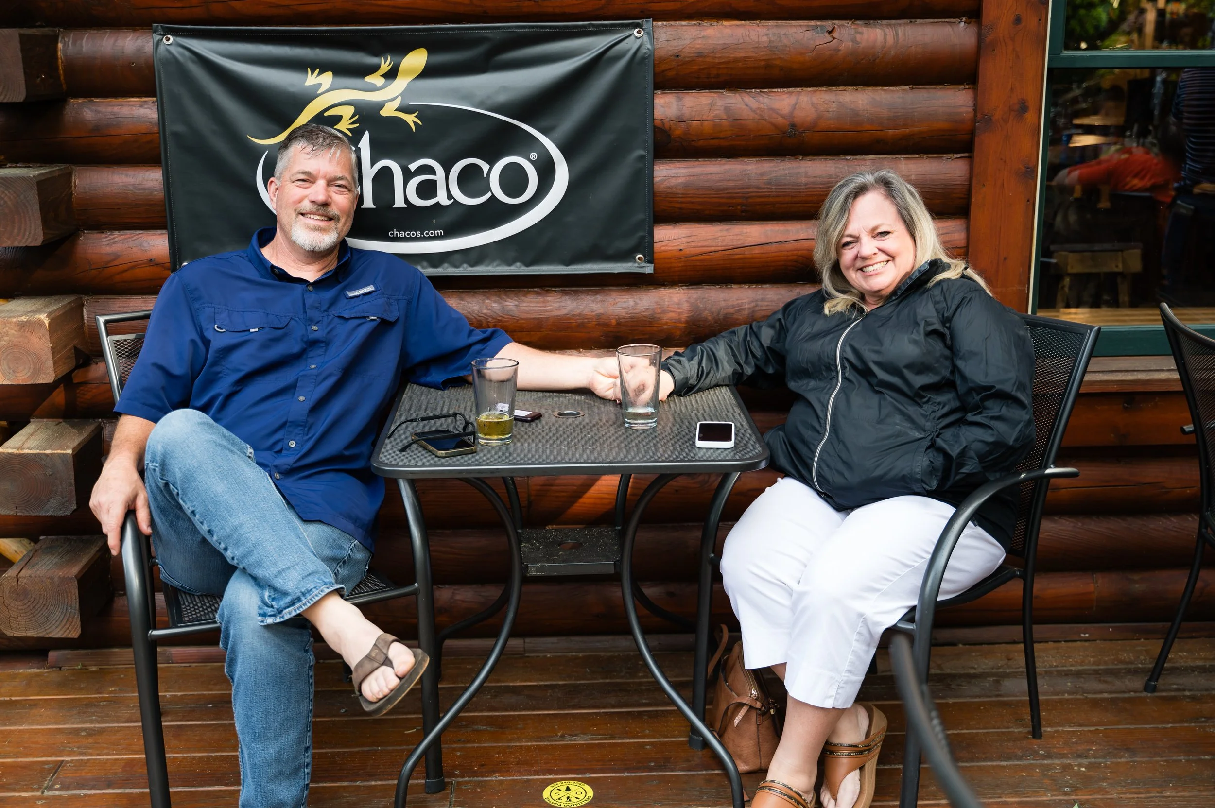A man and woman sitting at an outdoor table, smiling. The man is wearing a blue shirt and jeans, and the woman is wearing a black jacket and white pants. They are holding hands across the table. There are glasses of drinks in front of them and a mobi