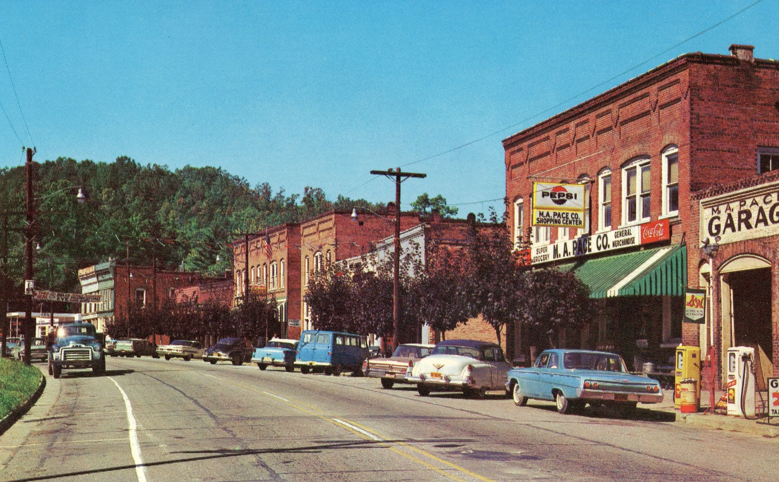 A small downtown street scene with vintage cars parked along the curb, brick buildings with signs, and a clear blue sky overhead.