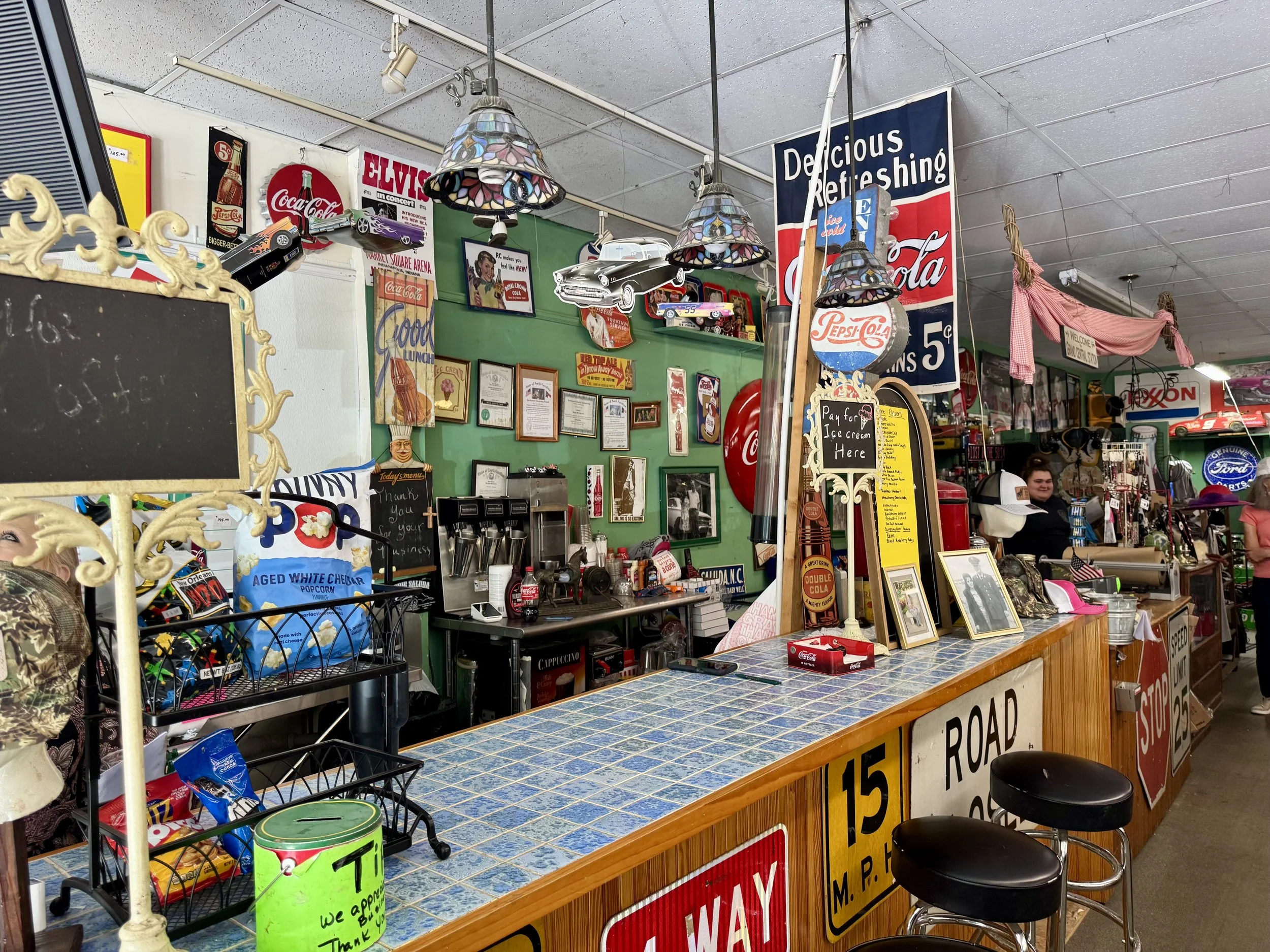 Interior of a vintage and thrift store with various nostalgic and retro signs, posters, and memorabilia. There are hanging Tiffany-style lamps above a tiled counter, and various items such as framed pictures and collectible signs are visible.