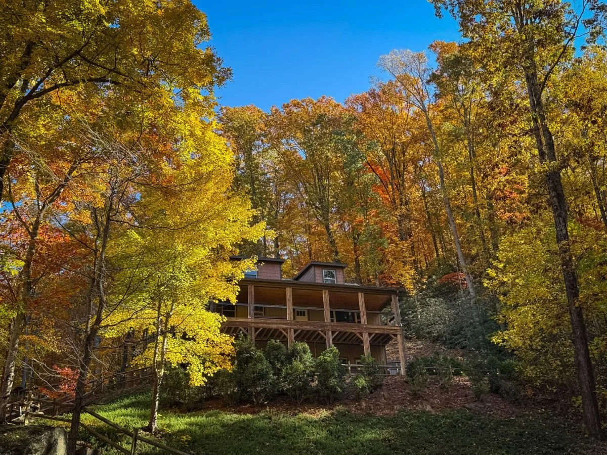 A house on a wooded hillside surrounded by fall-colored trees under a bright blue sky.