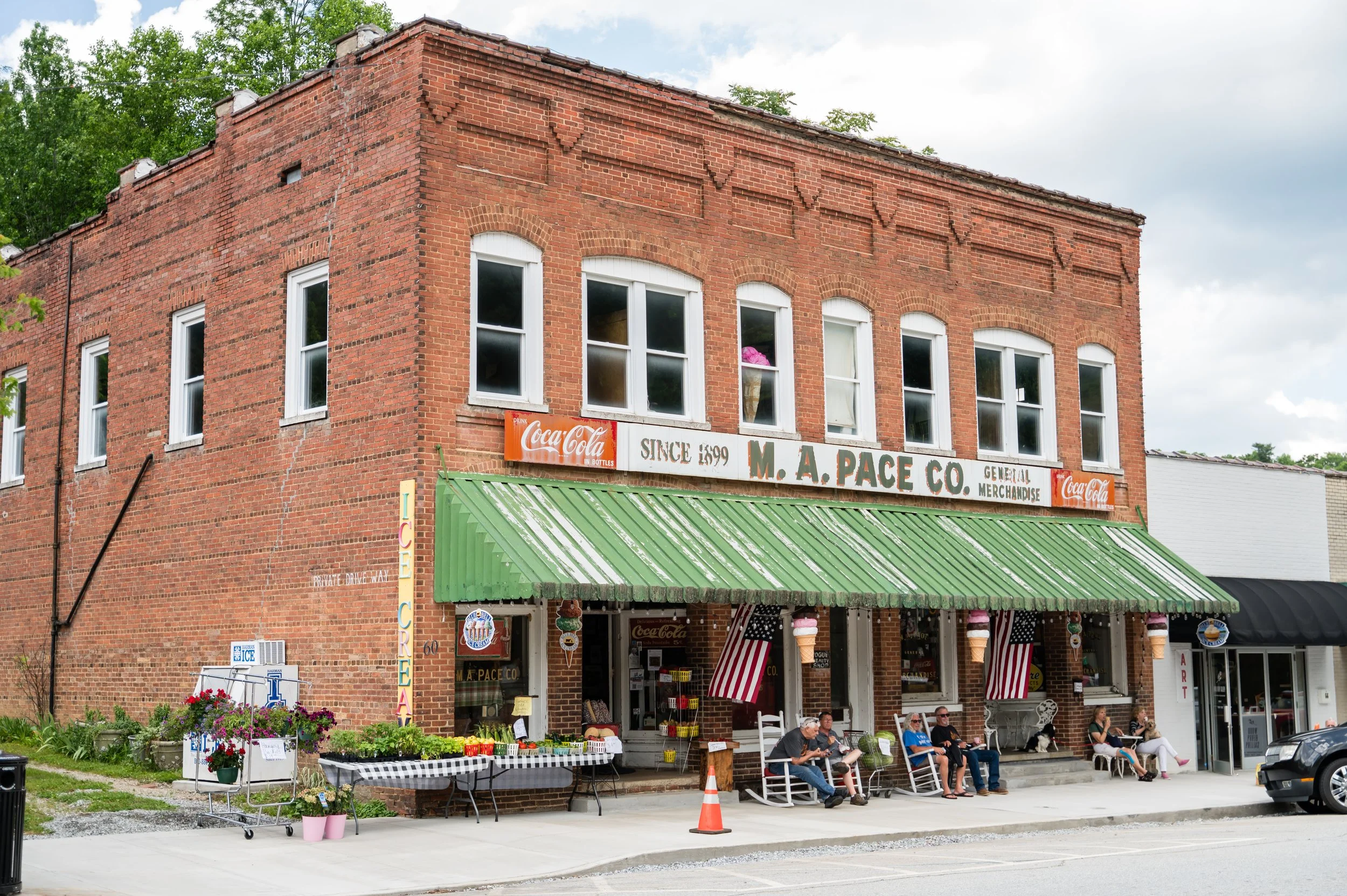 A historic brick building with a green awning, outdoor seating, and people sitting and walking in front. Storefront signs include 'M. A. Pace Co.' and Coca-Cola advertisements. Flower pots and small vendor tables are also visible.