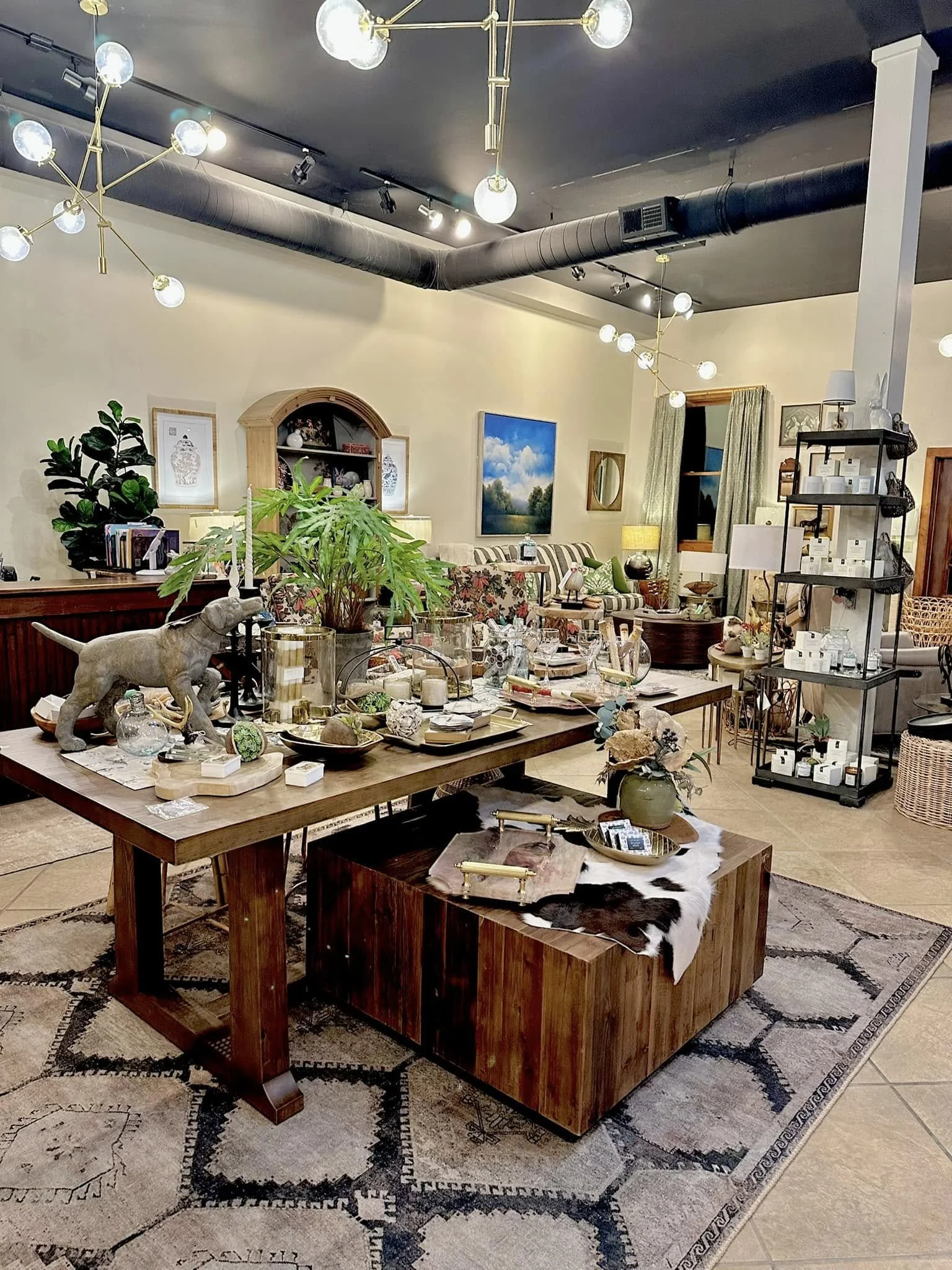A living room decorated with a mix of modern and rustic furniture, including a large wooden table with various decorative items, a striped sofa, plants, artwork on the walls, and modern lighting fixtures hanging from the ceiling.