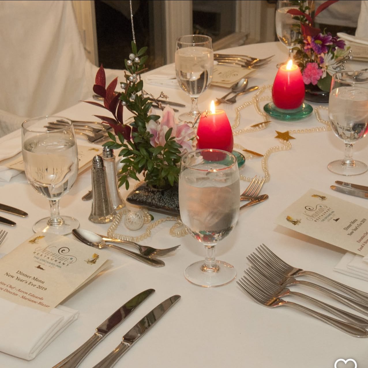 A decorated dining table with water glasses, silverware, red candles, floral arrangements, and menus, set for a festive dinner event.